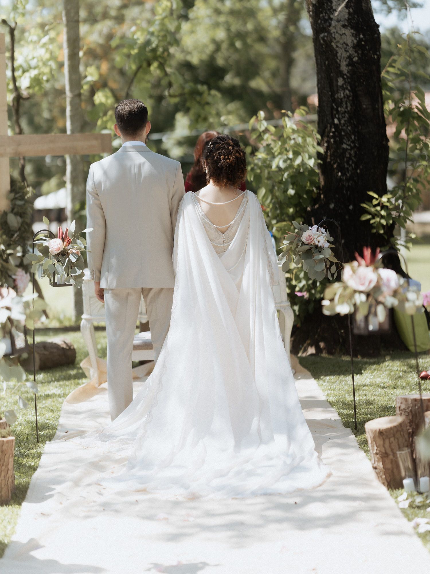 A couple in wedding attire stands on a white walkway surrounded by trees and white floral decorations in an outdoor garden.