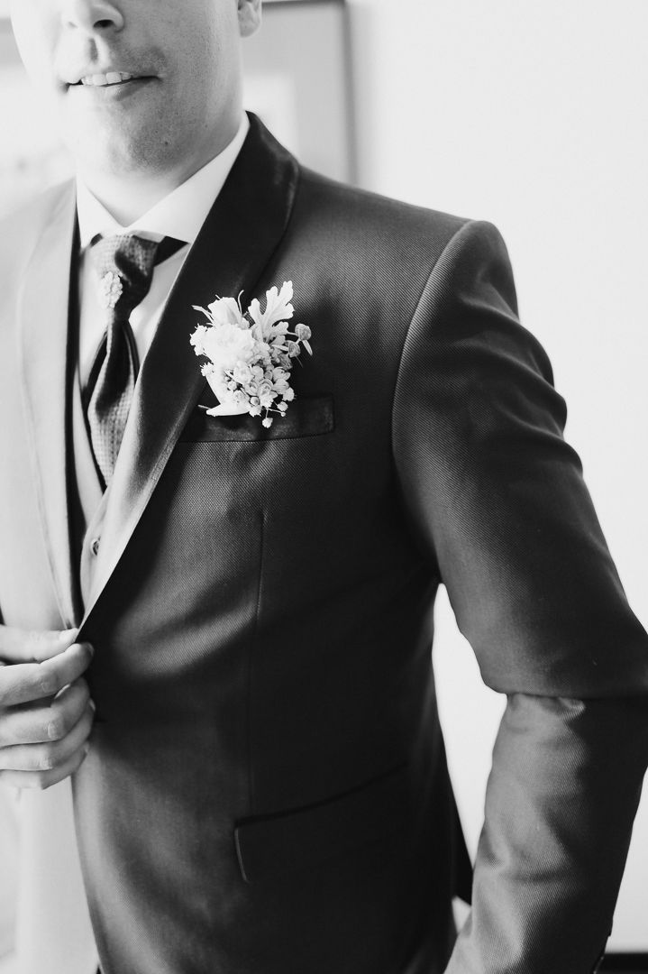 Close up black and white photo of formal attire showing a dark suit jacket with white floral boutonniere.