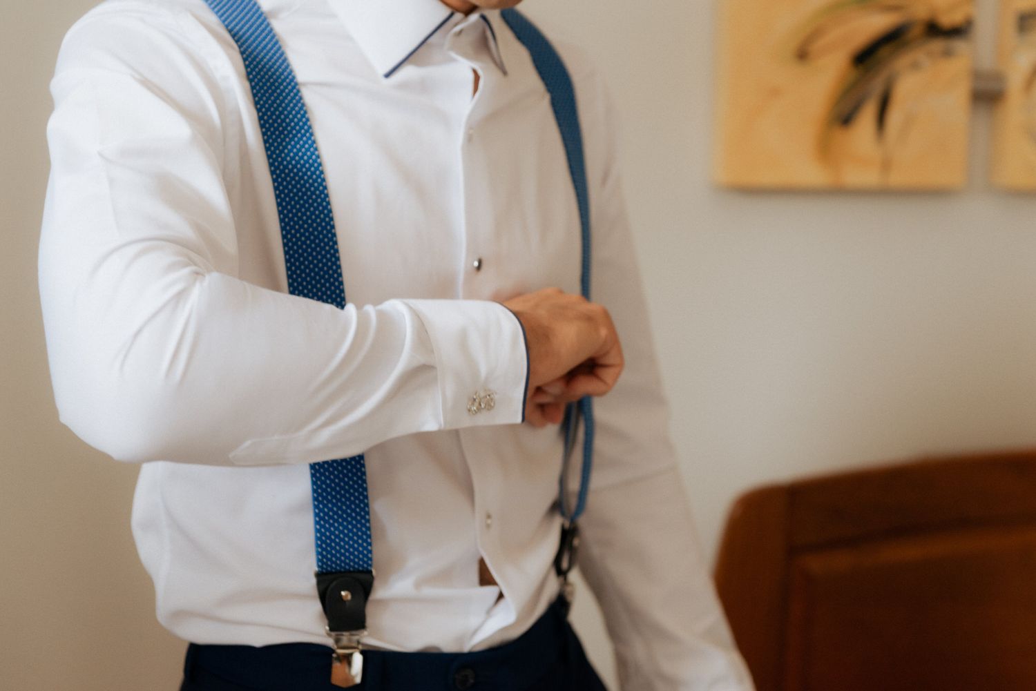 Person wearing crisp white dress shirt and navy blue suspenders while getting dressed for formal occasion.
