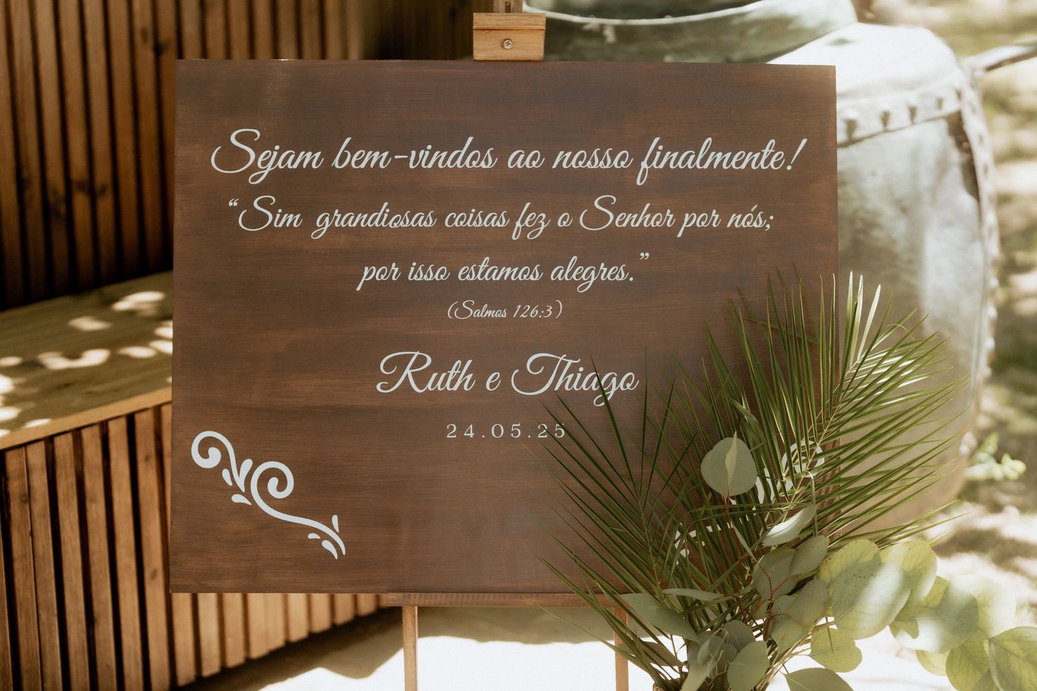 Wooden wedding sign with Portuguese text and palm frond decoration in natural lighting.