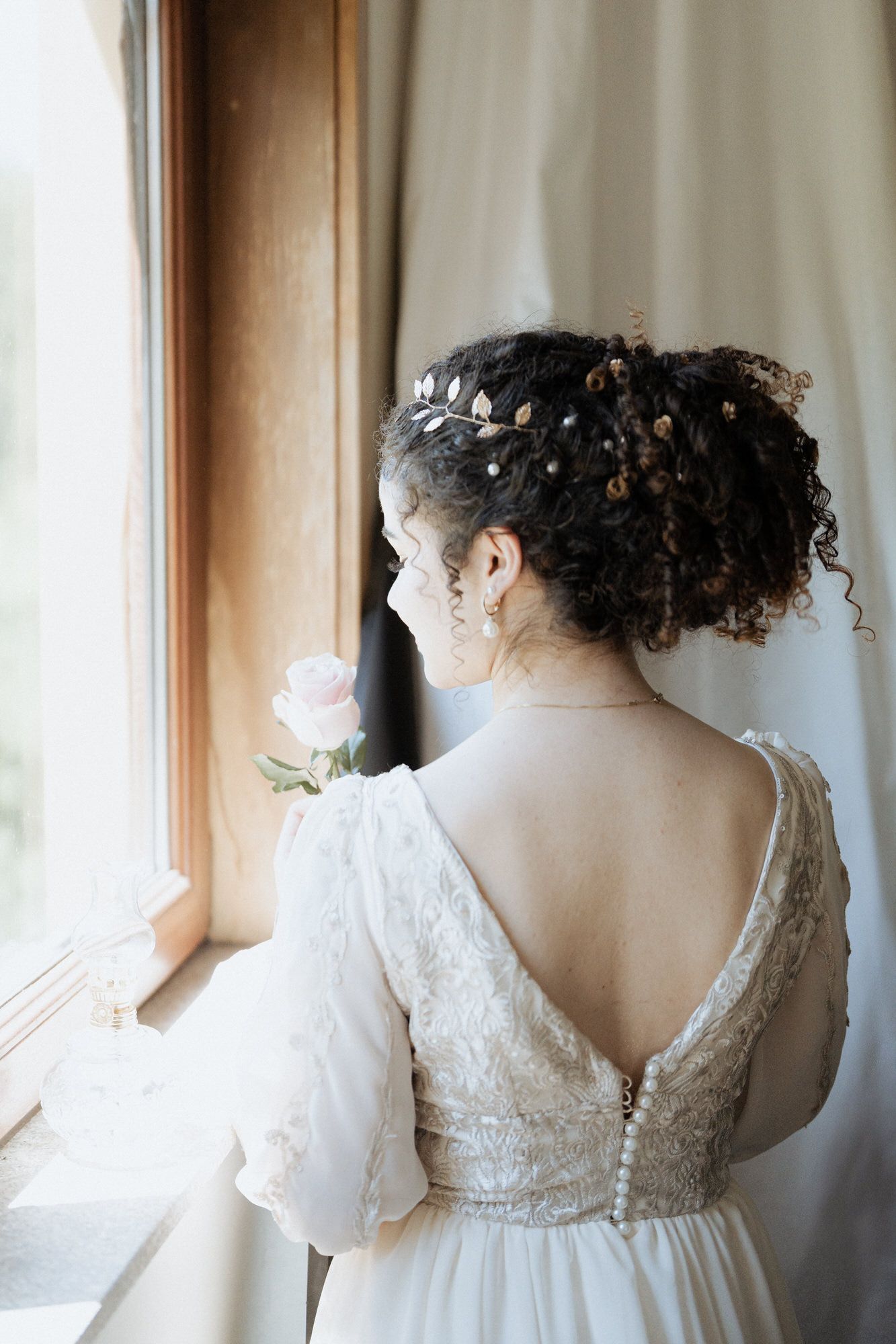 Back view of bride in vintage lace wedding dress with curly updo and crystal hair accessories near window.