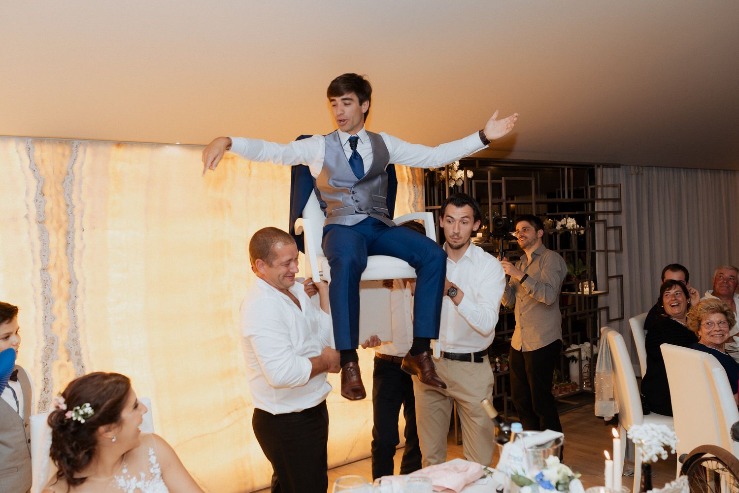Wedding guests lift and carry a person in a vest during a celebration at a reception venue with warm lighting and decor.