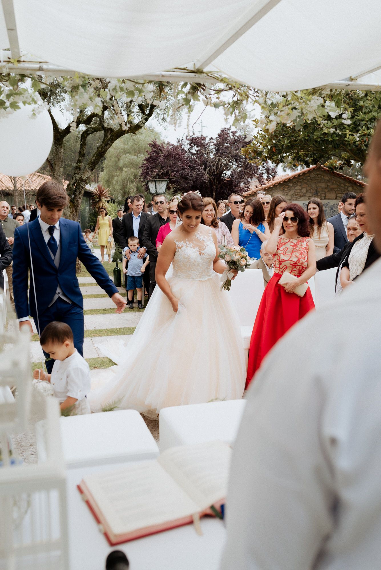 An outdoor wedding ceremony under a white canopy with guests gathered around a decorated aisle and altar.