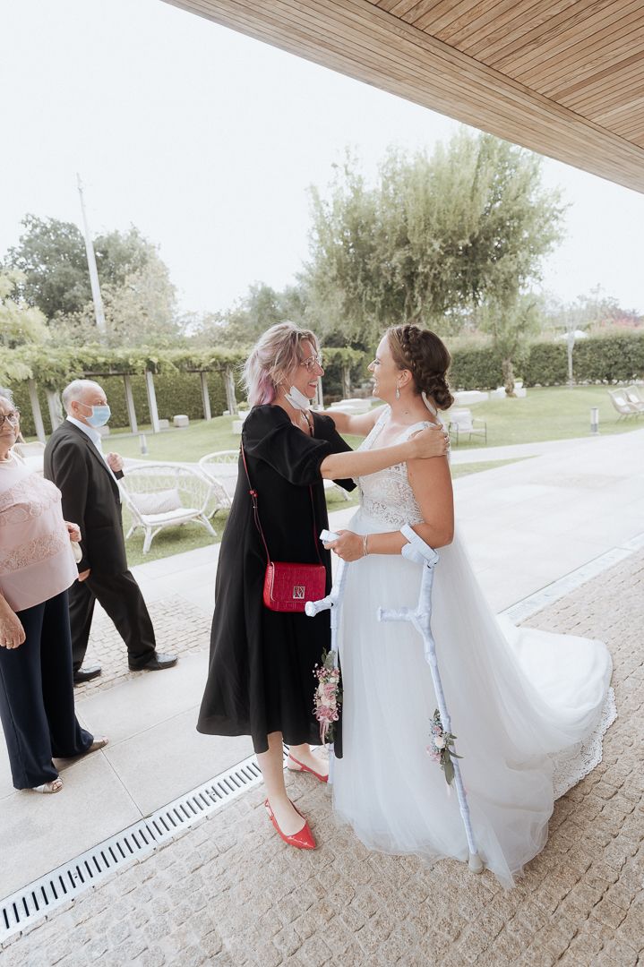 Two women embrace at an outdoor wedding venue, one wearing a flowing white gown and the other in an elegant black dress.
