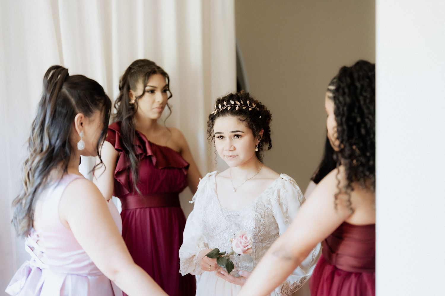Bridesmaids in burgundy dresses help adjusting the bride's lace gown in elegant wedding preparation moment.