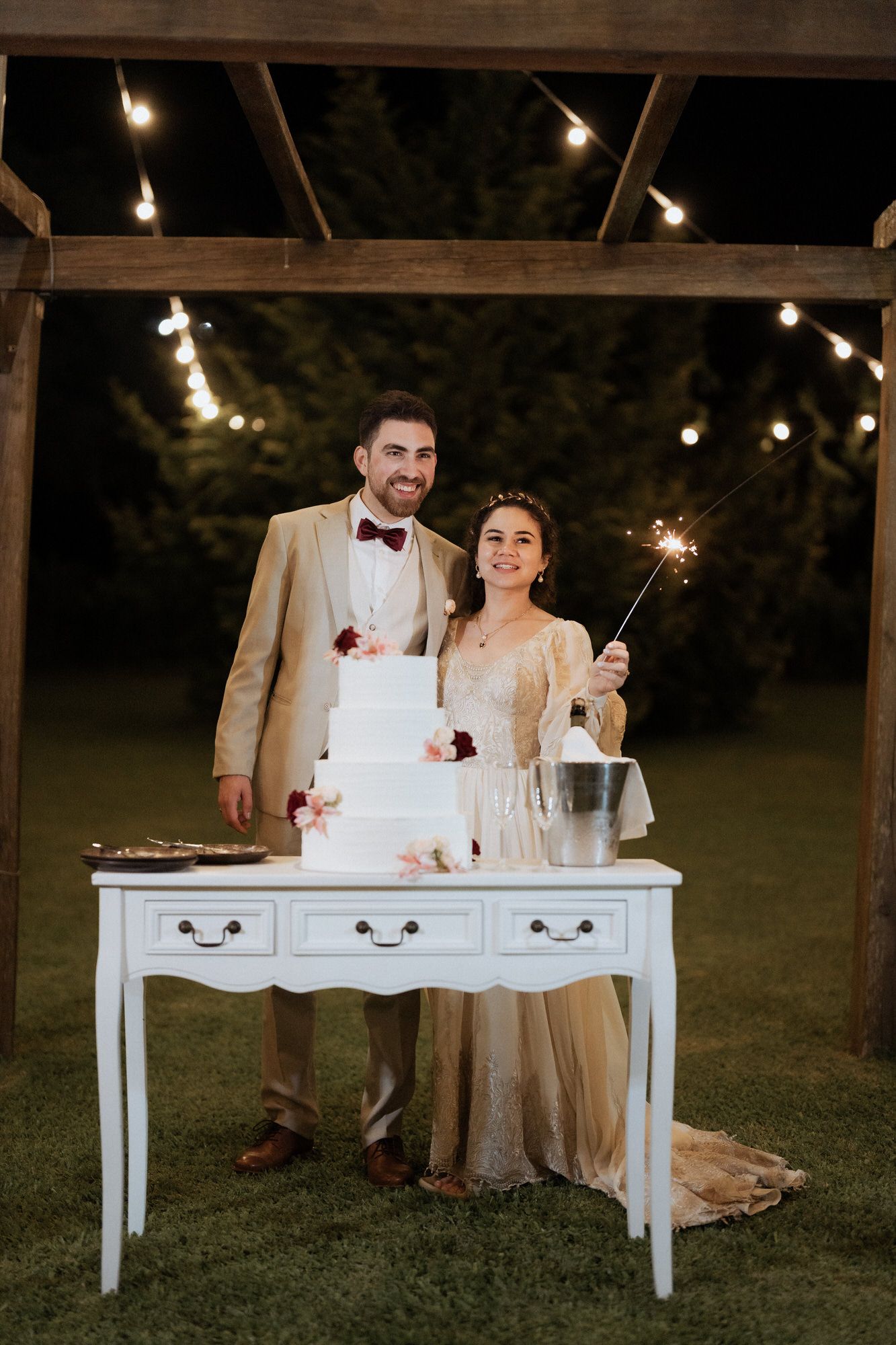 A couple stands at a white vintage console table with a wedding cake and sparklers under outdoor string lights at dusk.