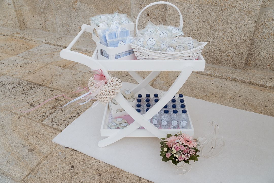 A vintage white serving cart decorated with pink flowers holds wedding glasses and accessories on a stone patio.