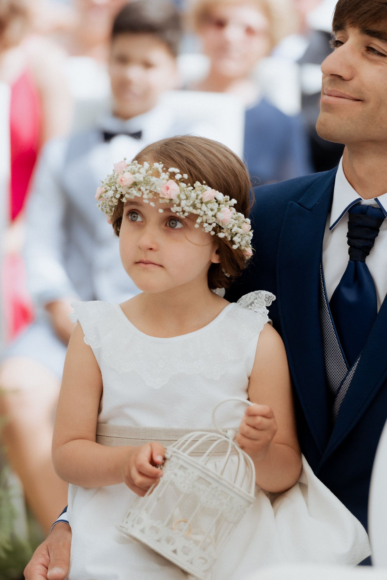 A flower girl in a white dress holds a decorative birdcage during a wedding ceremony.