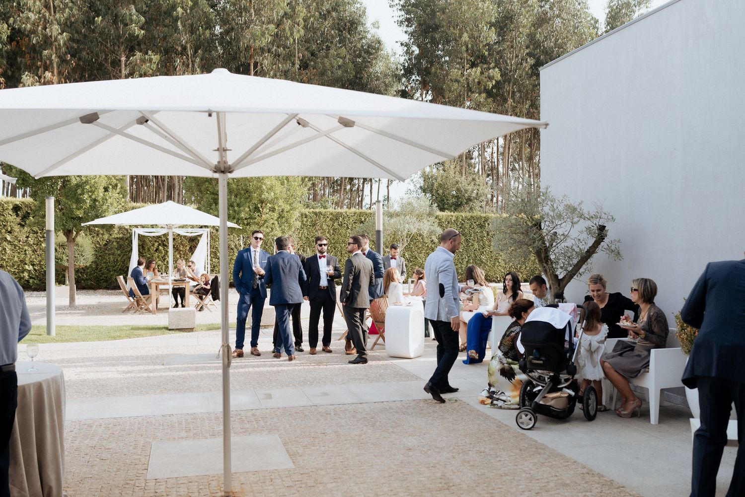 Guests mingle under white umbrellas at an elegant outdoor event venue with olive trees in the background.