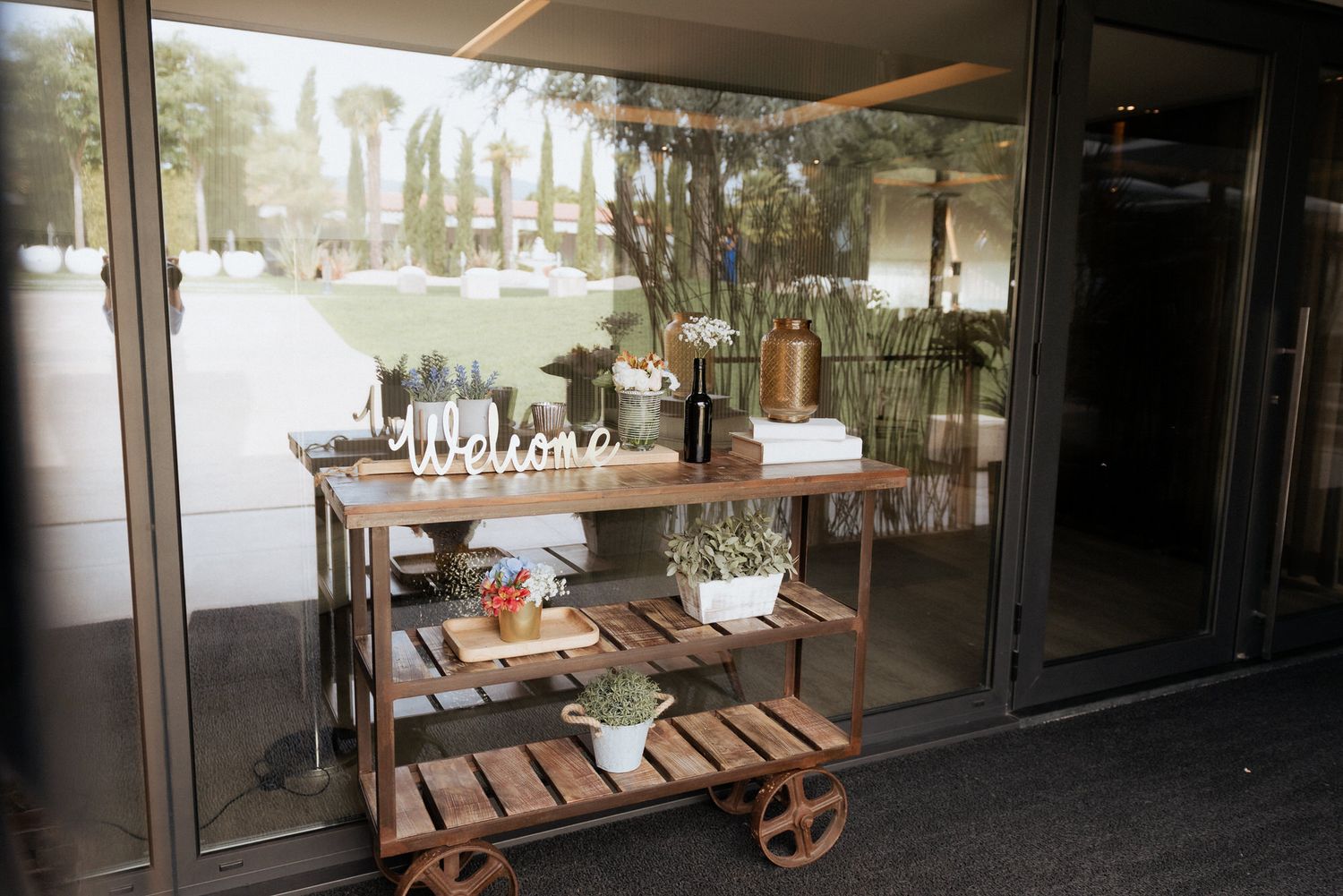 A rustic wooden plant cart with potted plants displayed behind glass windows at a garden shop entrance.