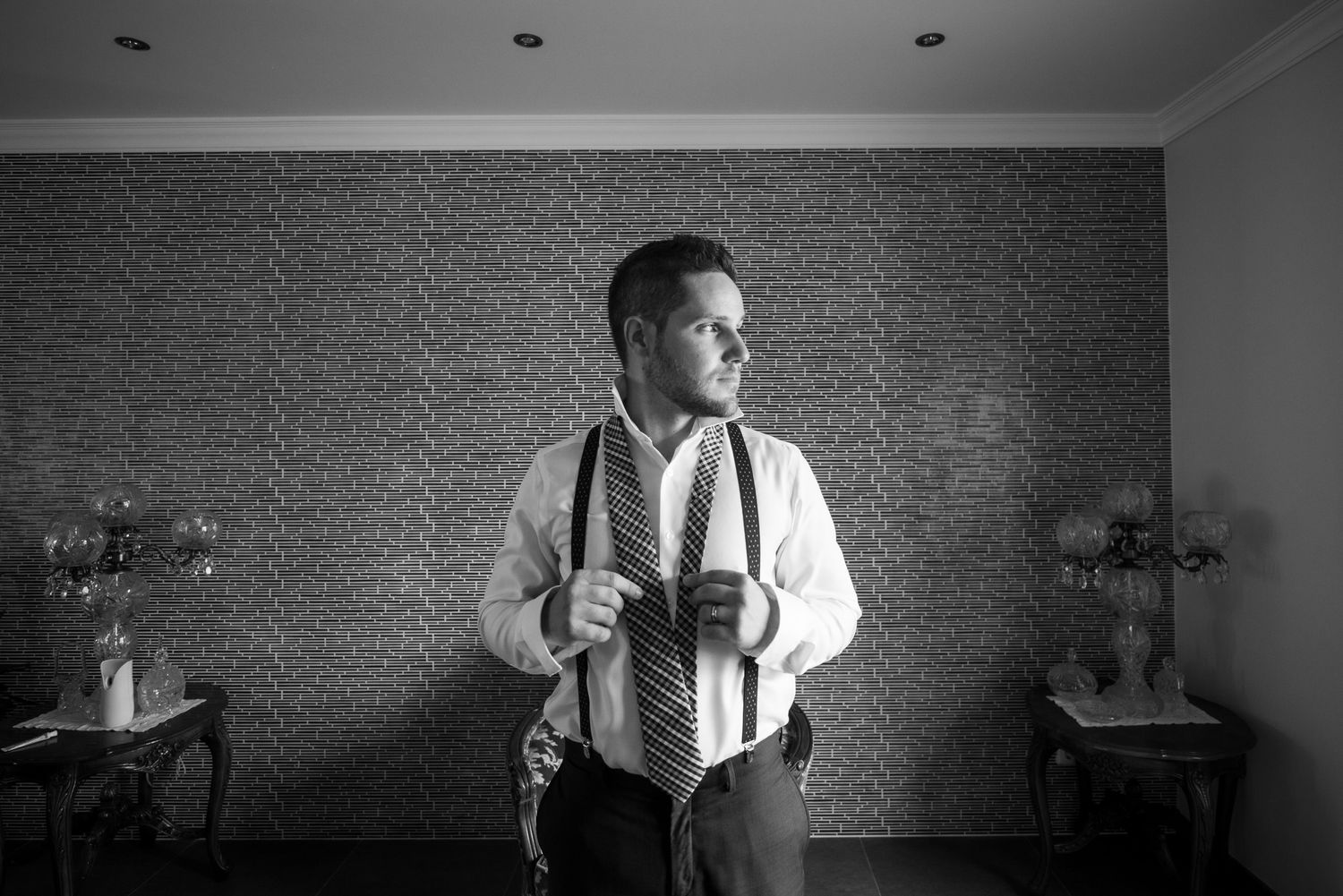 Um retrato monocromático contra uma parede de tijolos texturizados mostrando traje formal do noivo com suspensórios.