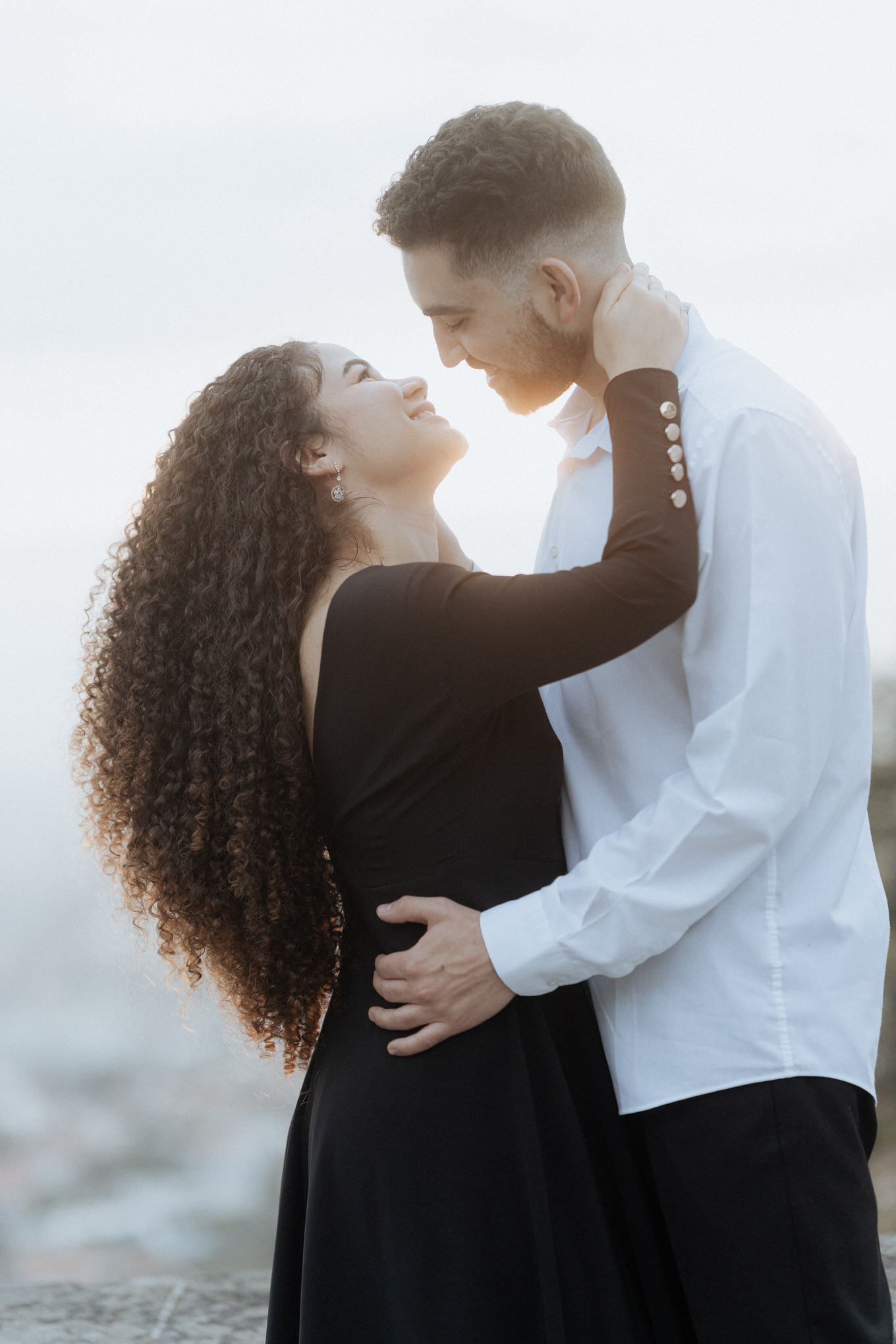 Casal romântico em trajes preto e branco se abraça contra o céu suavemente iluminado ao pôr do sol.