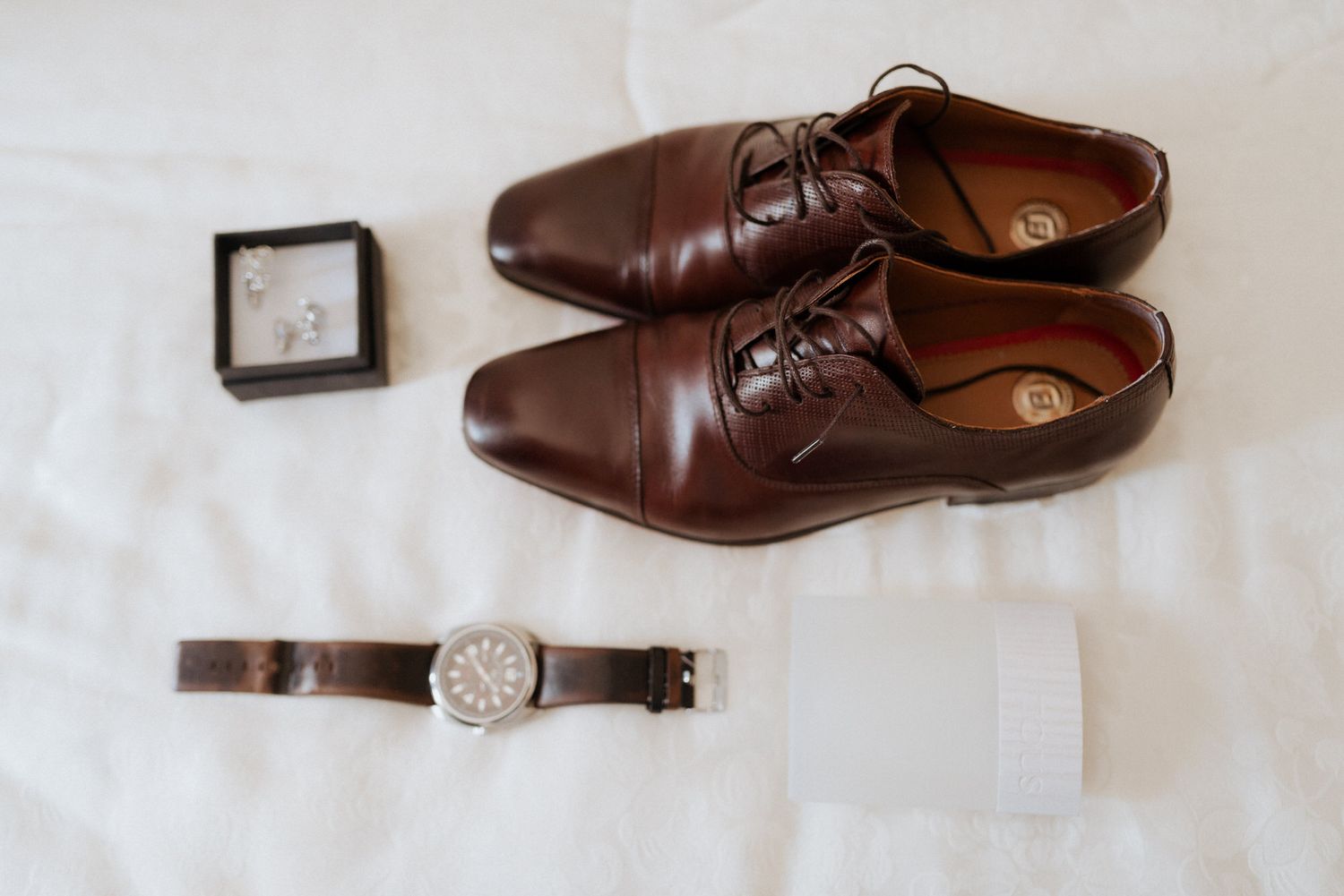 Brown leather dress shoes paired with a classic wristwatch and small clock on white linen surface.