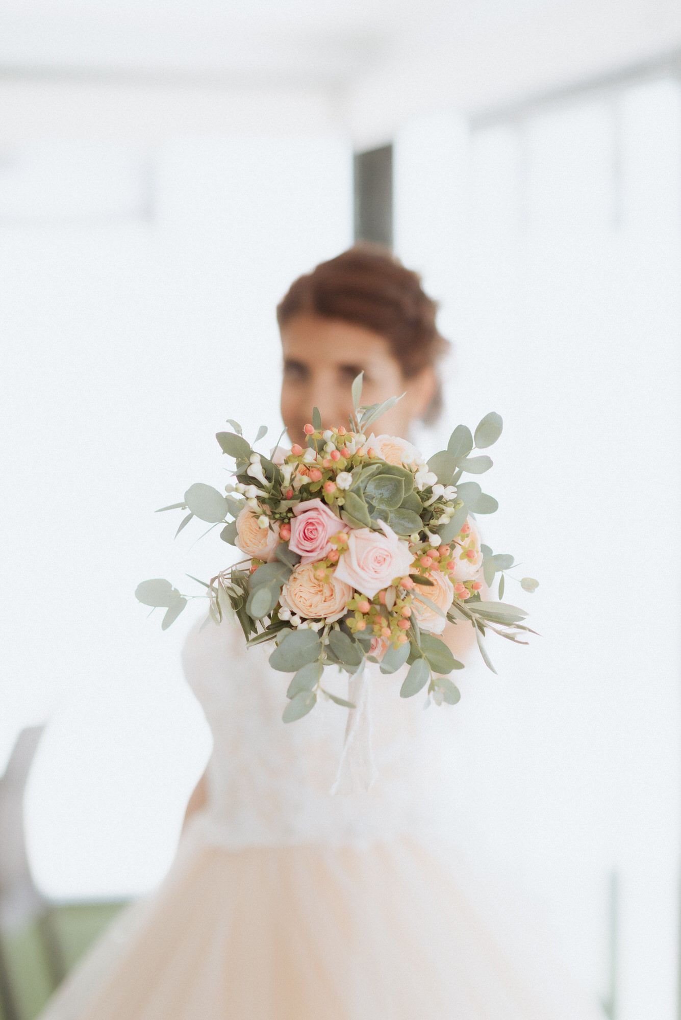 Delicate bridal bouquet with blush roses and eucalyptus held up in front of white wedding dress in soft focus.