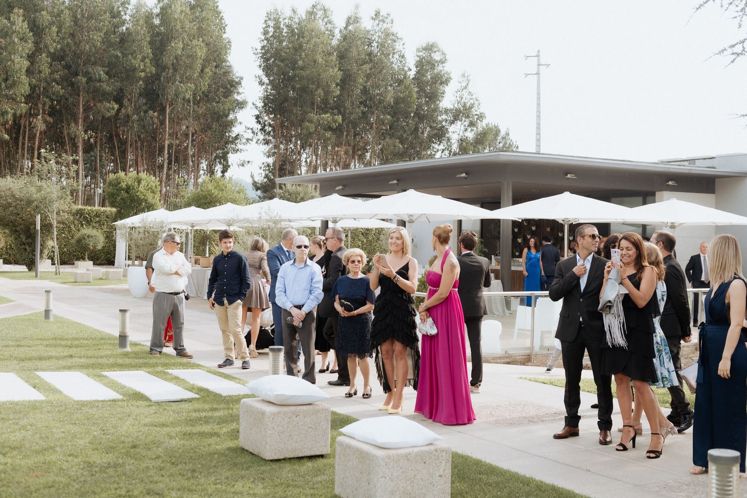 Wedding guests gather outside a modern venue with white canopies and trees in the background on a sunny afternoon.