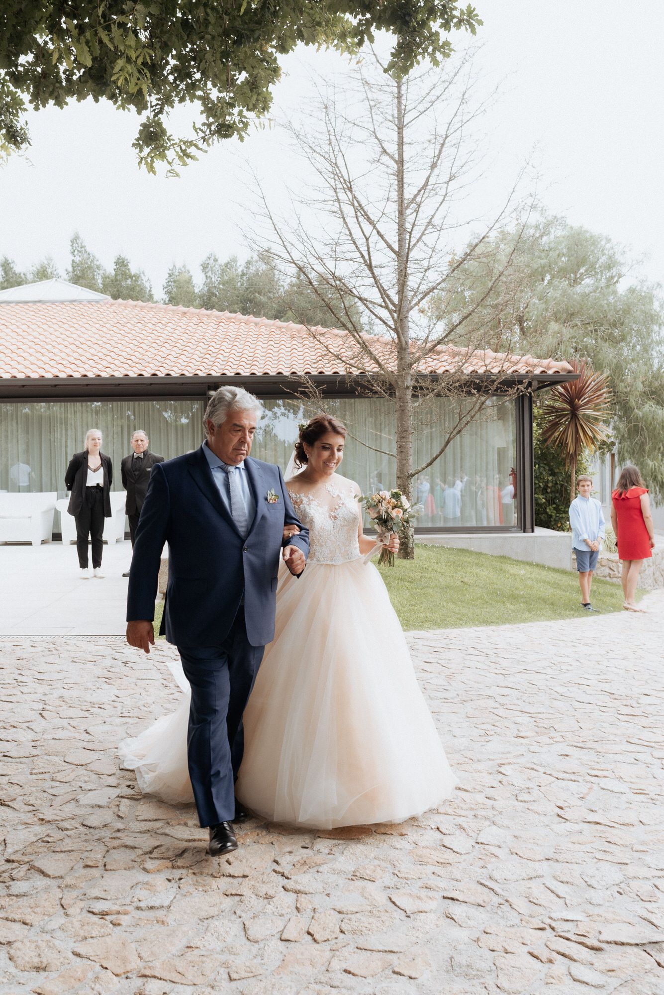 A bride in a white ballgown walks along a stone pathway outside a Spanish-style building with guests in the background.