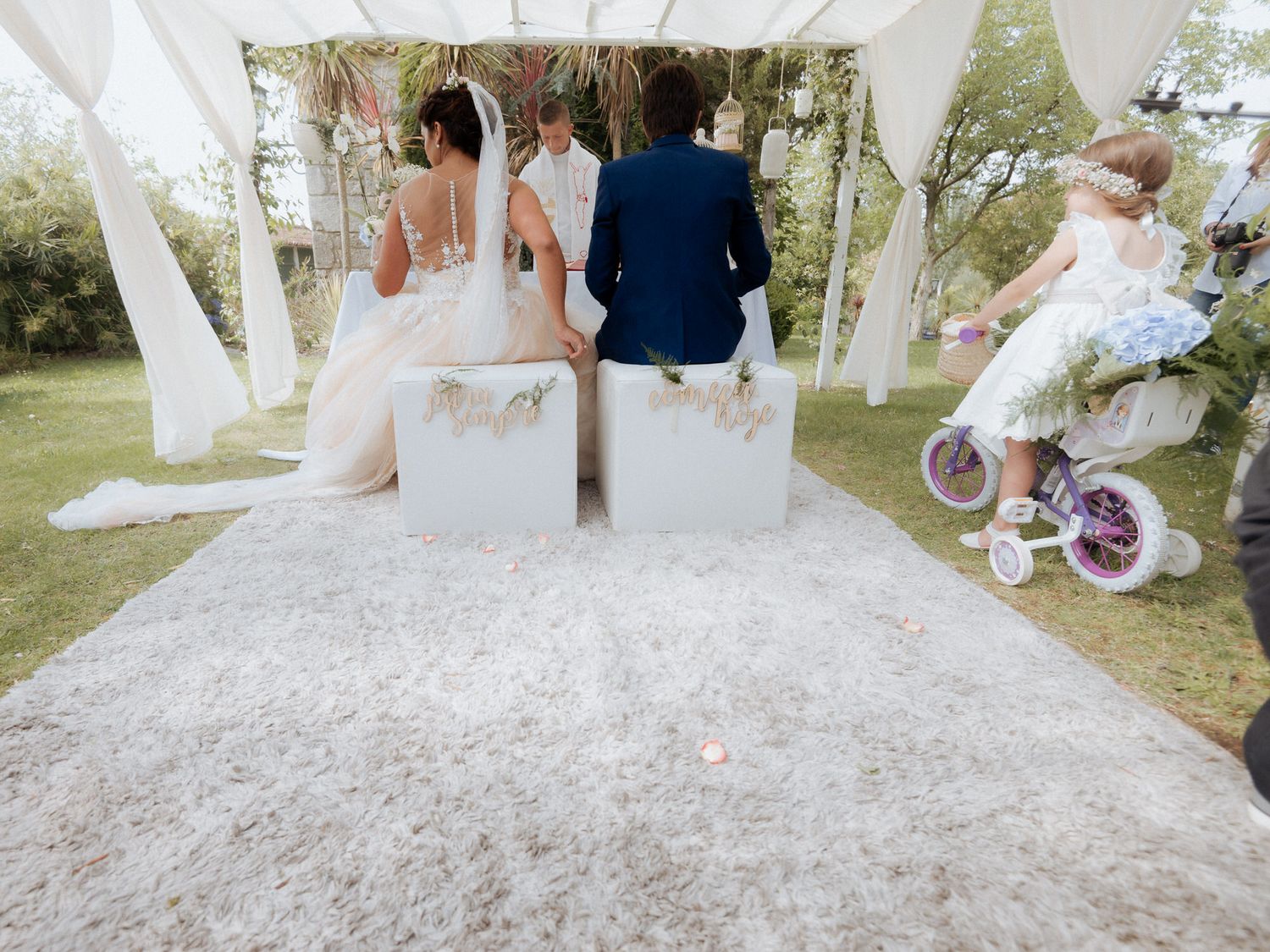 Wedding ceremony guests seated on white benches under a floral arch in an outdoor garden setting.