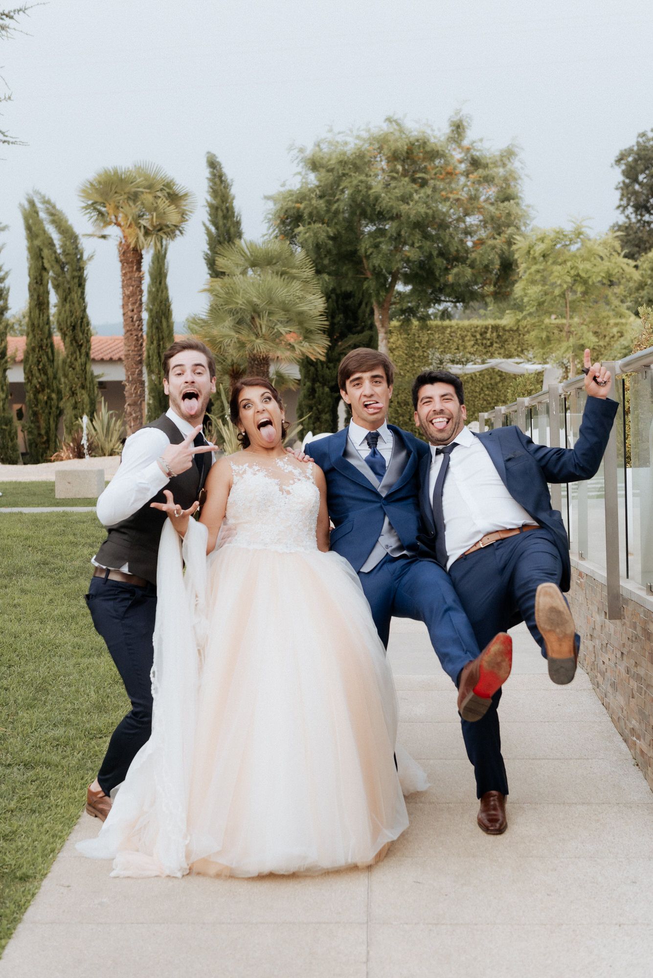 Wedding party members strike energetic poses on a garden pathway lined with cypress trees and palm trees.