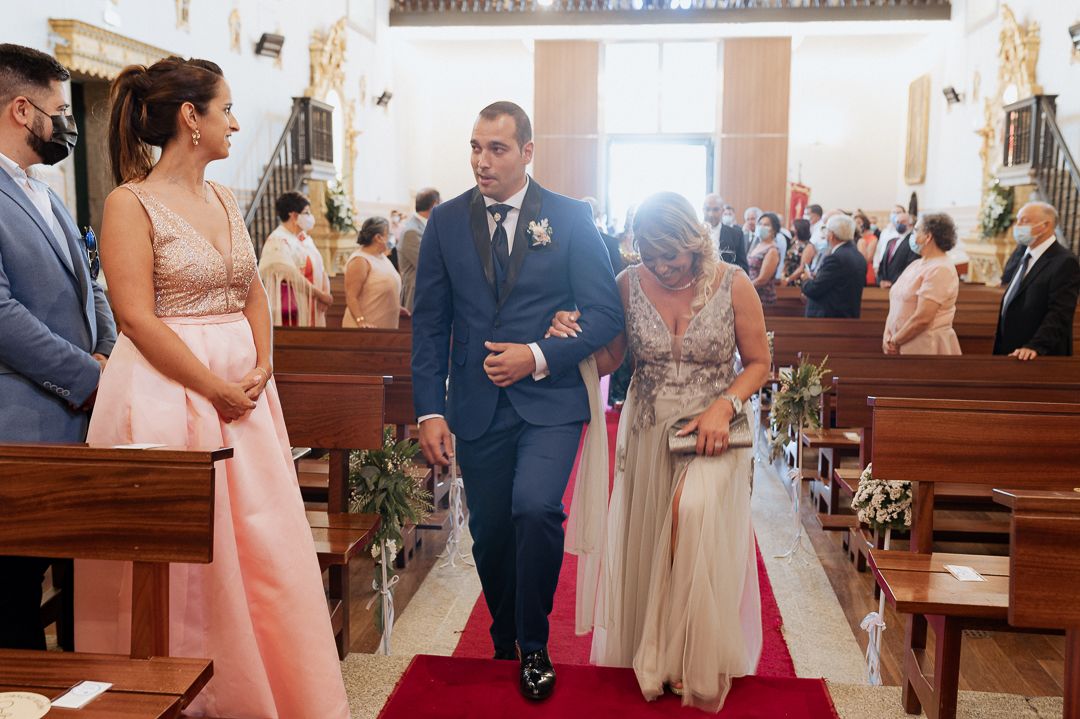 Wedding guests in formal attire gather inside a traditional church with wooden pews and a red carpet aisle.
