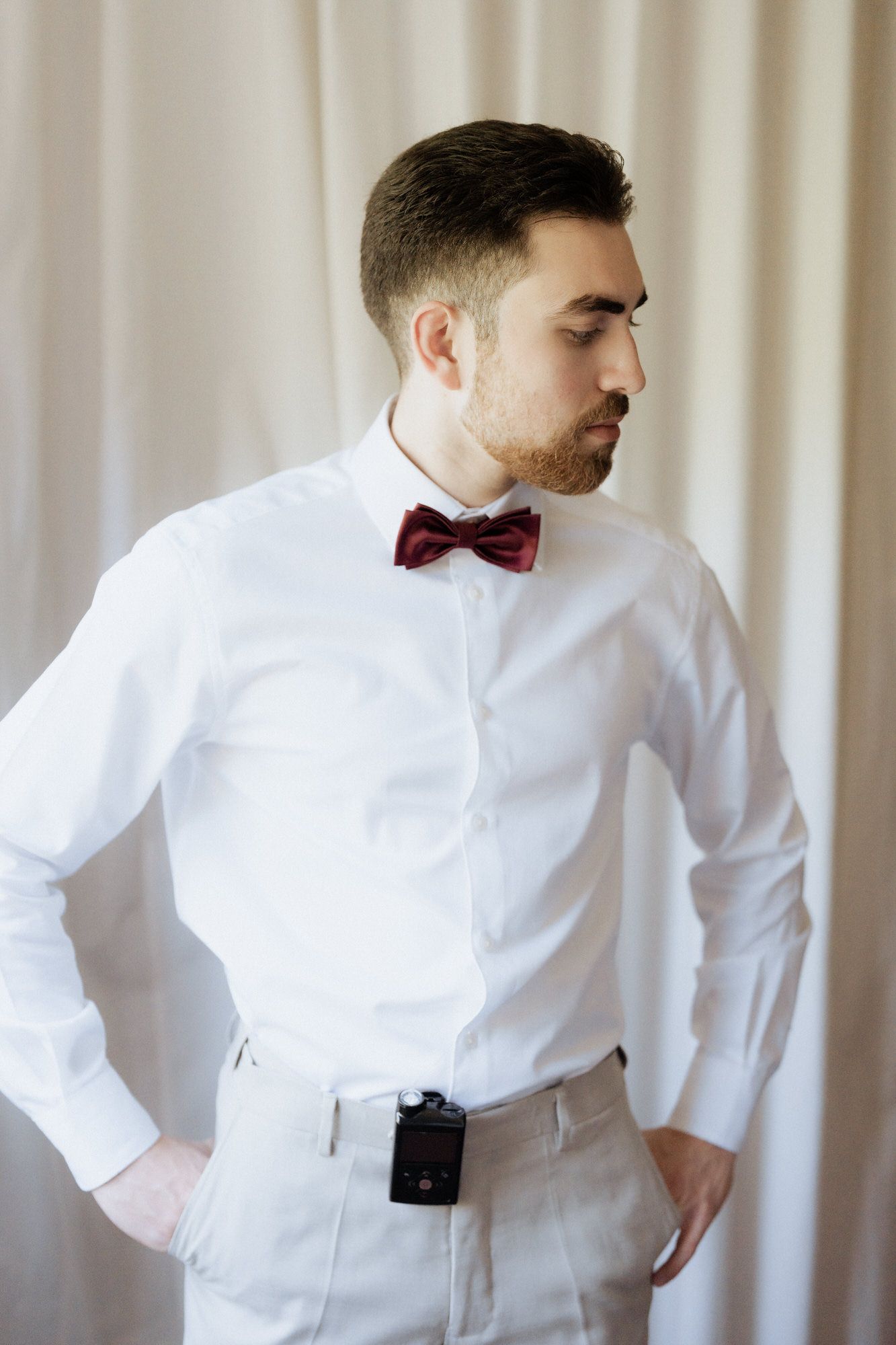 A person in a crisp white dress shirt and burgundy bow tie poses while looking to the side in formal attire.