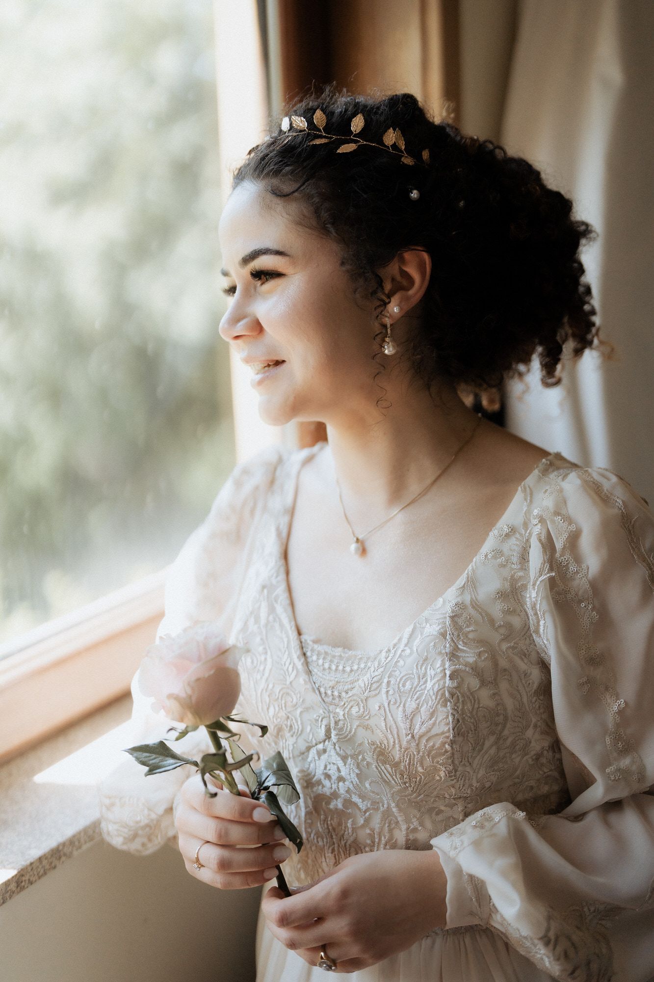 A person in a white lace wedding dress gazes out a window while holding a small bouquet of flowers.