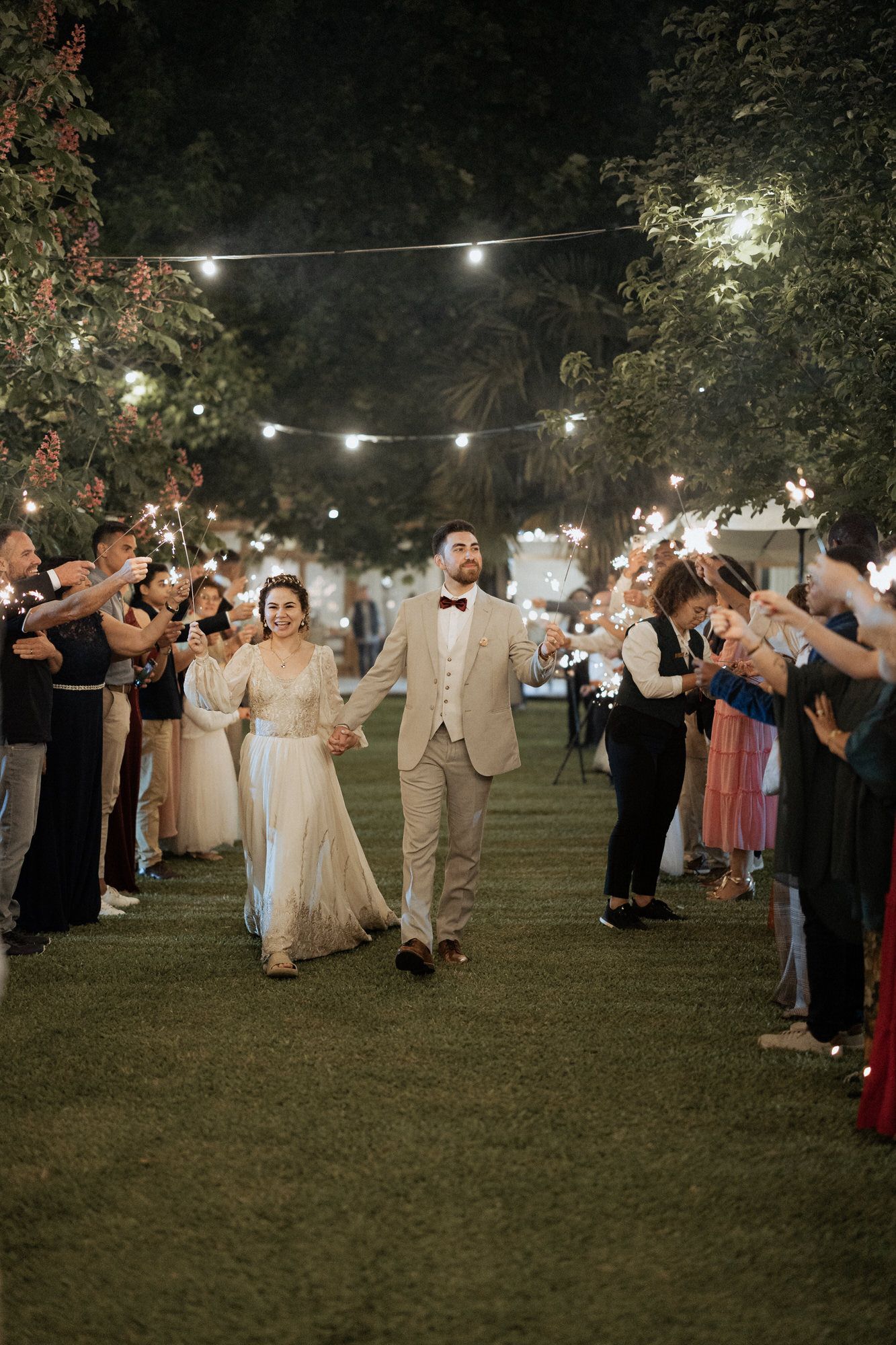 Newlyweds walk down an illuminated outdoor aisle at night while guests hold sparklers creating a magical exit.
