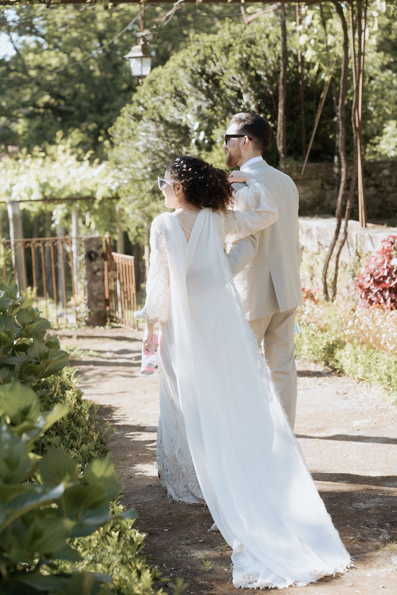 A couple in elegant white wedding attire walking along a garden path on a sunny day.