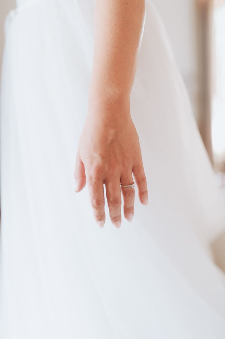 A delicate hand wearing a silver wedding ring against the soft white fabric of a wedding dress.