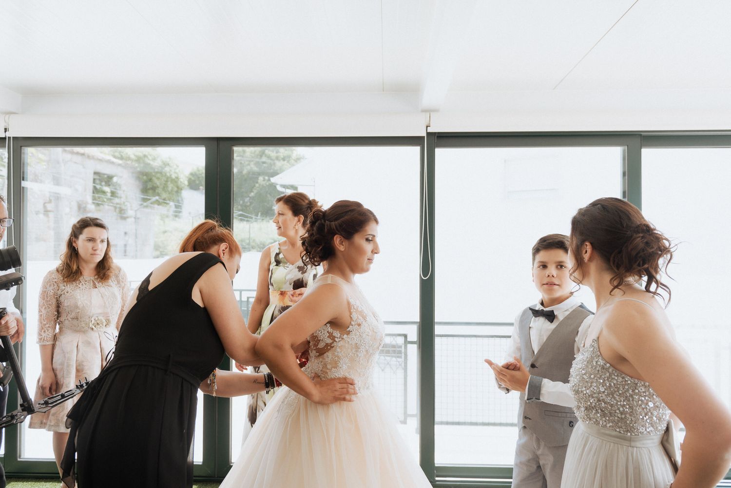 Wedding party members helping adjust a bride's dress inside a bright modern venue with large windows.