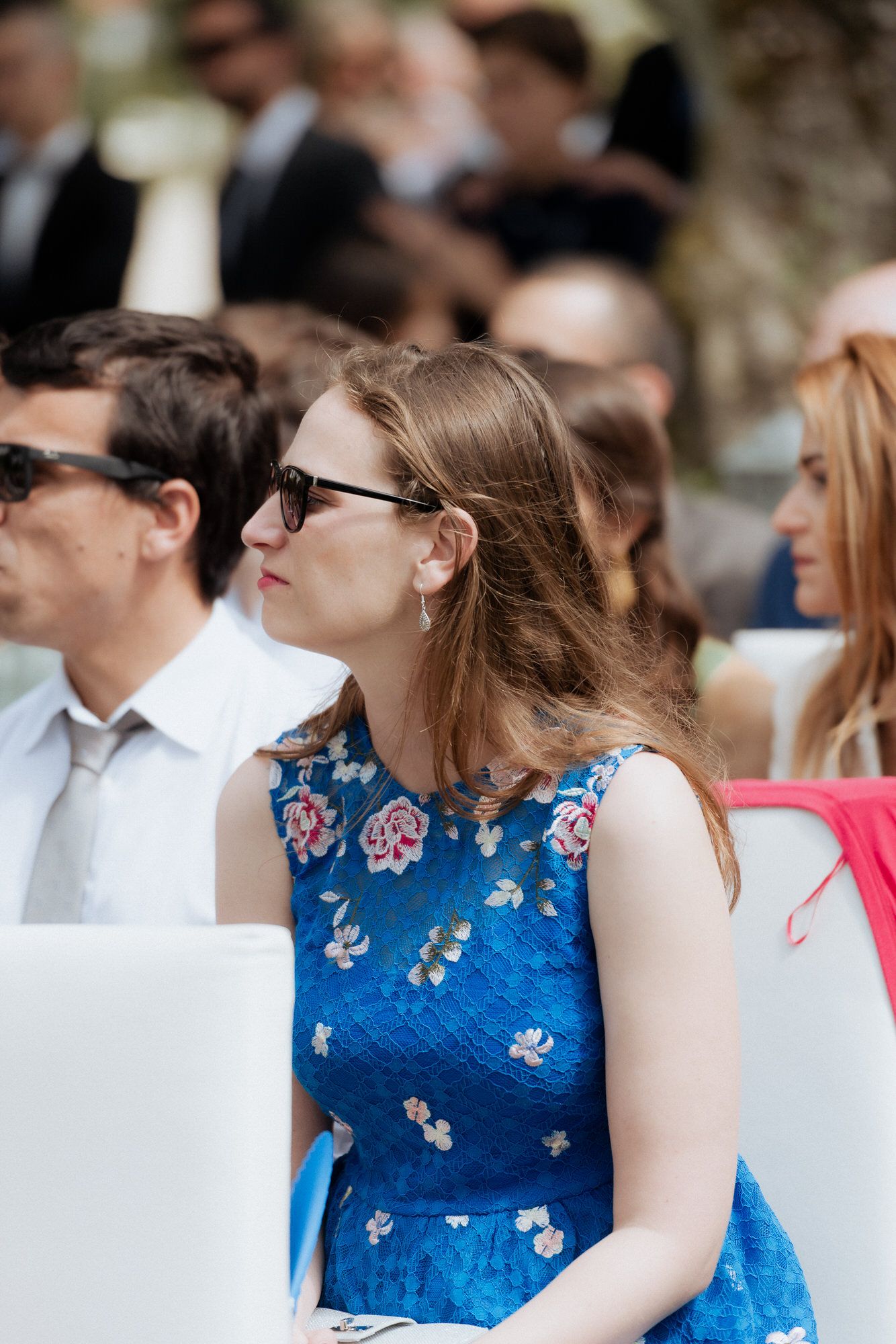 Woman in blue floral dress smiles while sitting outdoors at social gathering.