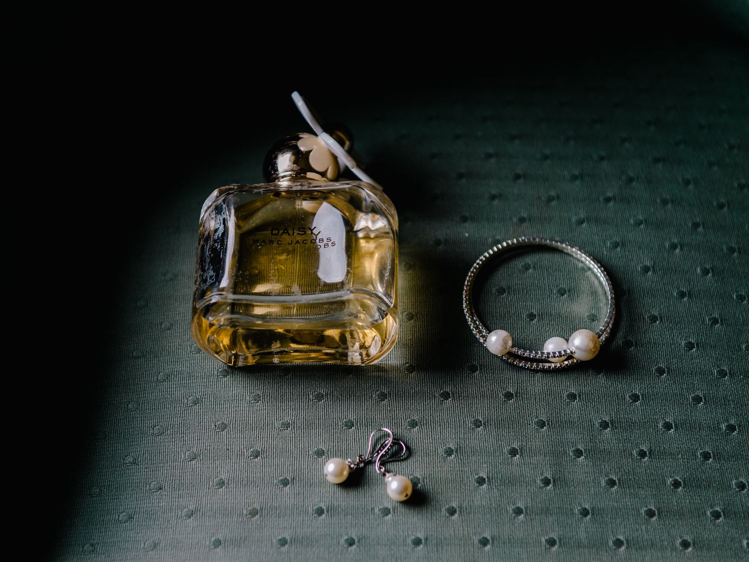 Acessórios de casamento, incluindo um frasco de perfume, anel de diamante e brincos de pérola em superfície texturizada escura.
