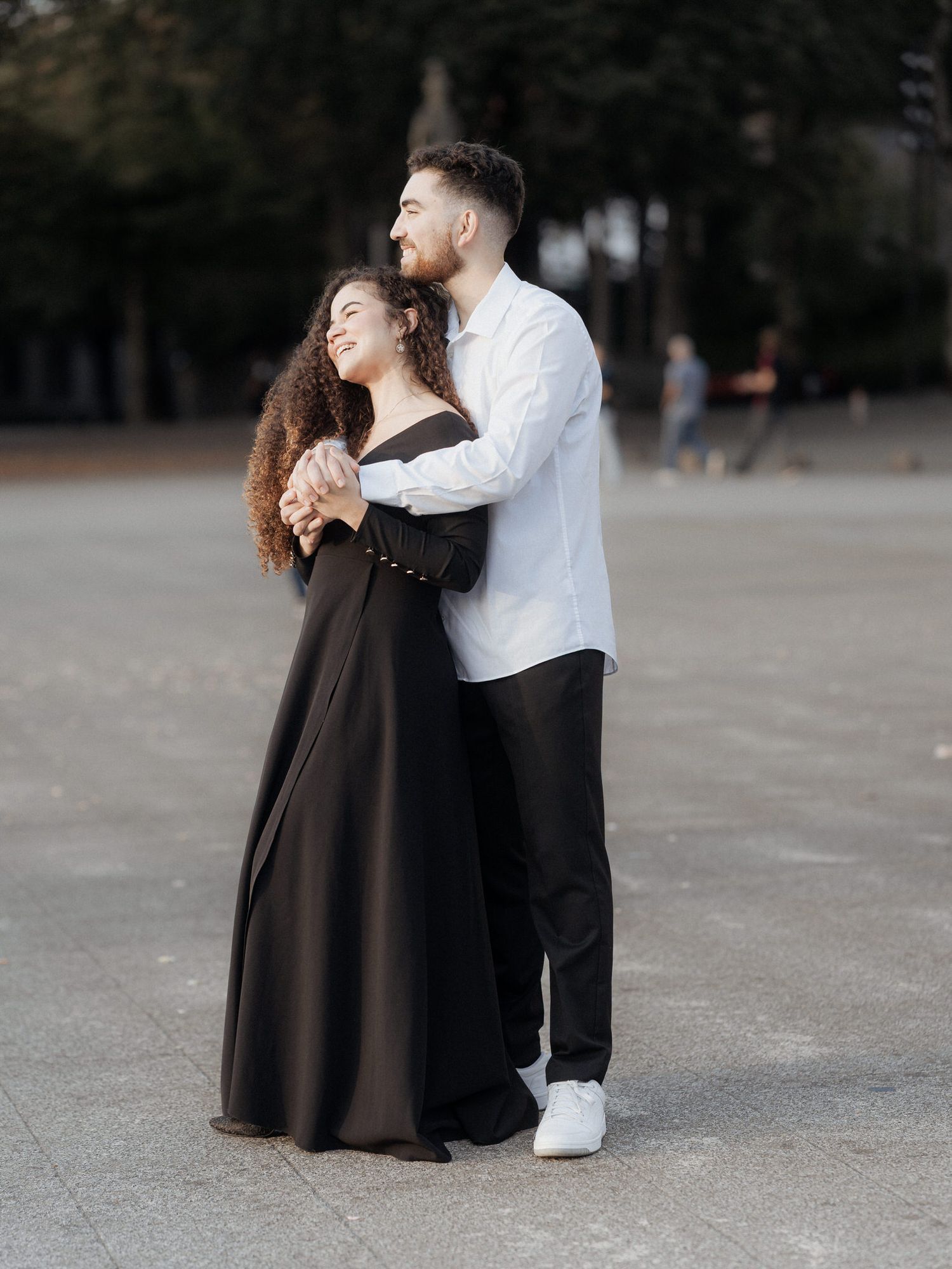 Um casal romântico se abraça ao ar livre, ela com um vestido preto esvoaçante e ele com uma camisa branca e calças escuras.