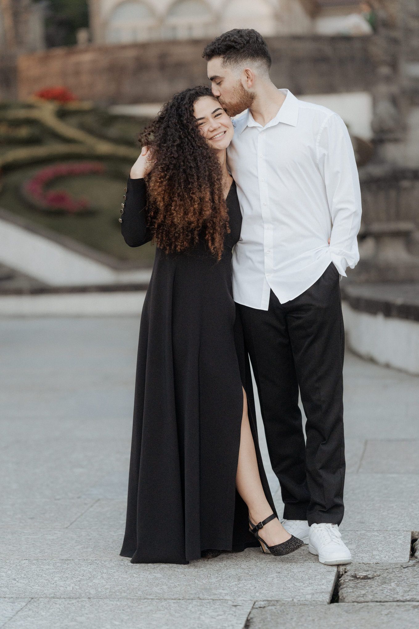 Stylish couple sharing tender moment outdoors, woman in long black dress with curly hair and man in white shirt.