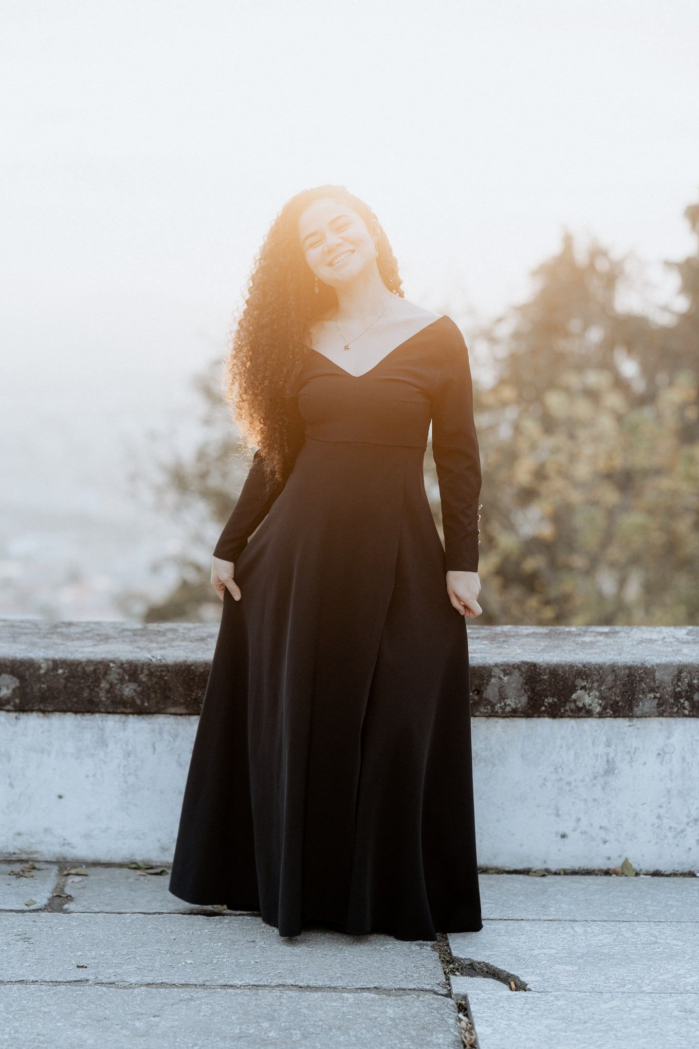Uma pessoa com um longo vestido preto está de pé contra um fundo enevoado, com seus cabelos cacheados refletindo a luz do sol.