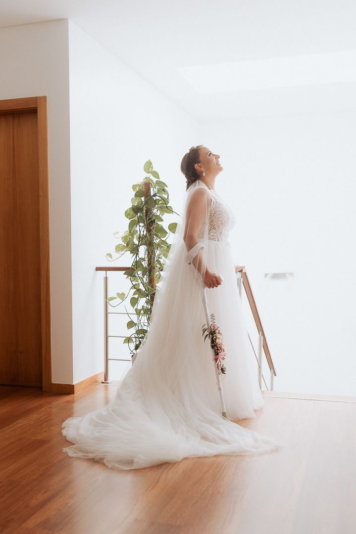A bride in a flowing white lace wedding gown stands near a wooden door and potted plant in natural lighting.