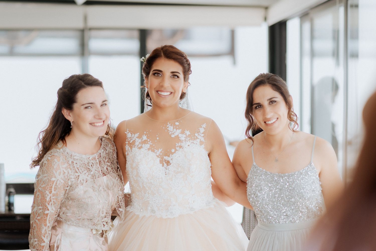 Three women in elegant formal wear smile together at a wedding celebration in a bright indoor setting.