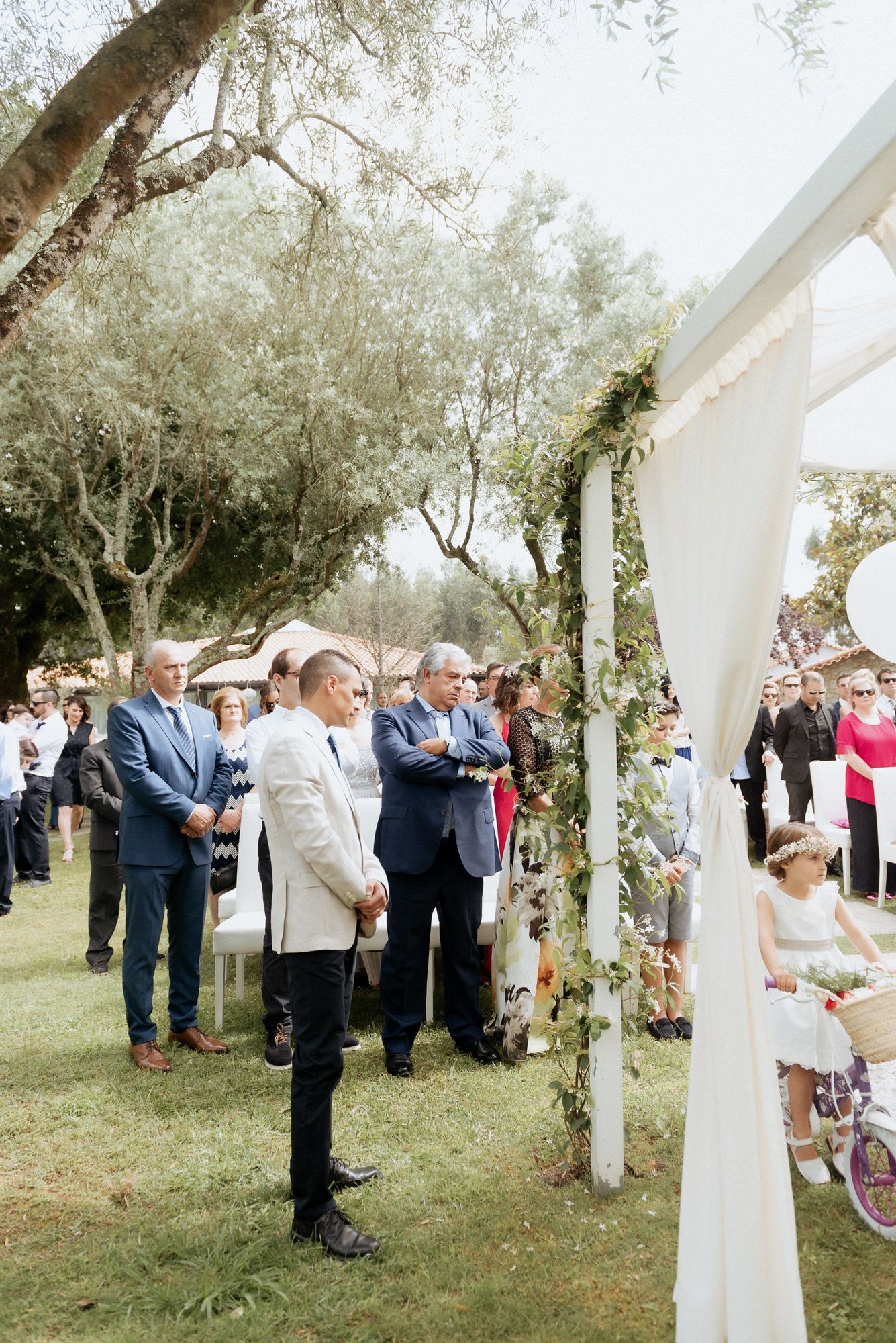 Outdoor wedding ceremony setup with white arch and guests gathered on grassy lawn under trees.