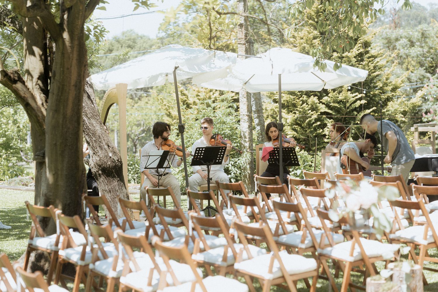 String quartet performs outdoors under white umbrellas with rows of empty wooden folding chairs in a garden setting.