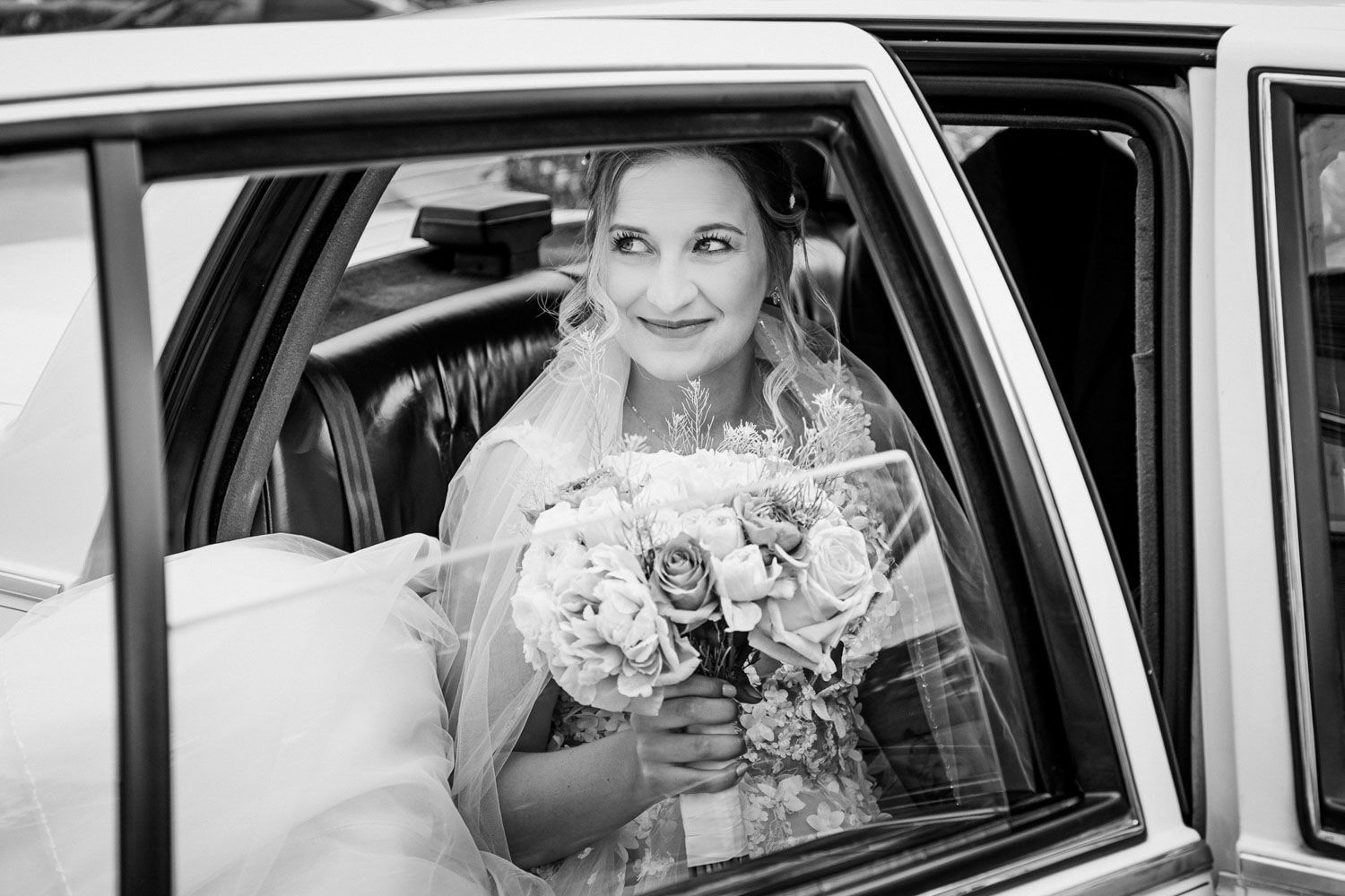 Egy klasszikus fekete-fehér fénykép, amelyen a menyasszony egy virágcsokrot tart, miközben egy vintage autóban ül.