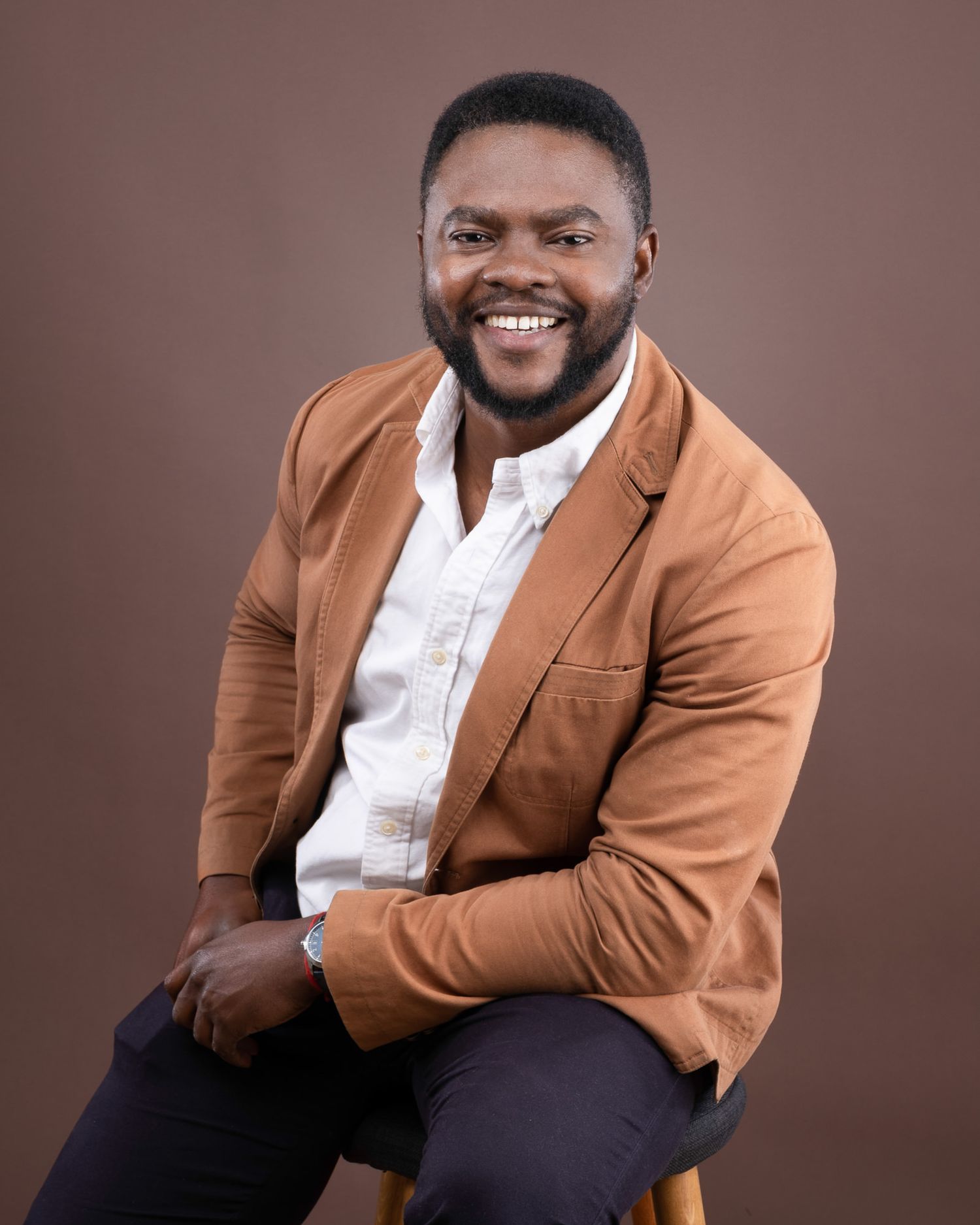Professional headshot of someone in a brown blazer and white shirt smiling warmly against a brown background.