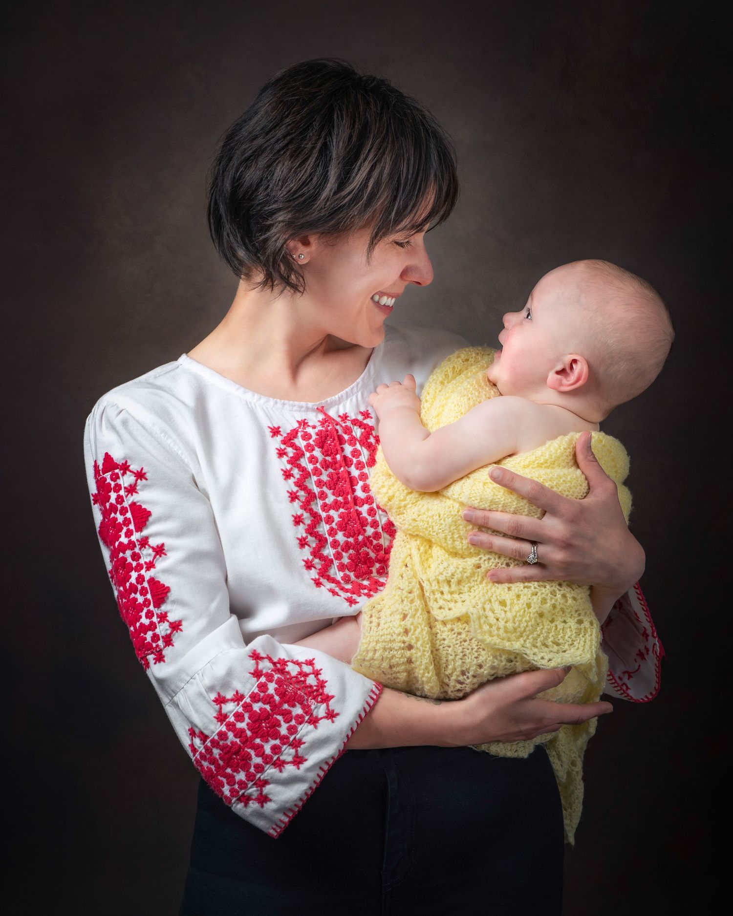 A tender moment shows someone in an embroidered white and red top holding a baby wearing yellow against a dark background.