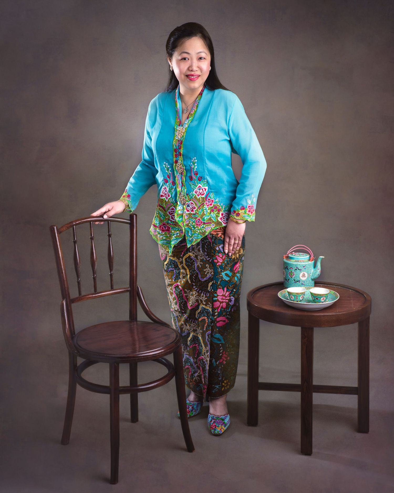 Person wearing turquoise cardigan and floral sarong stands between wooden chair and side table with teal teapot setup.
