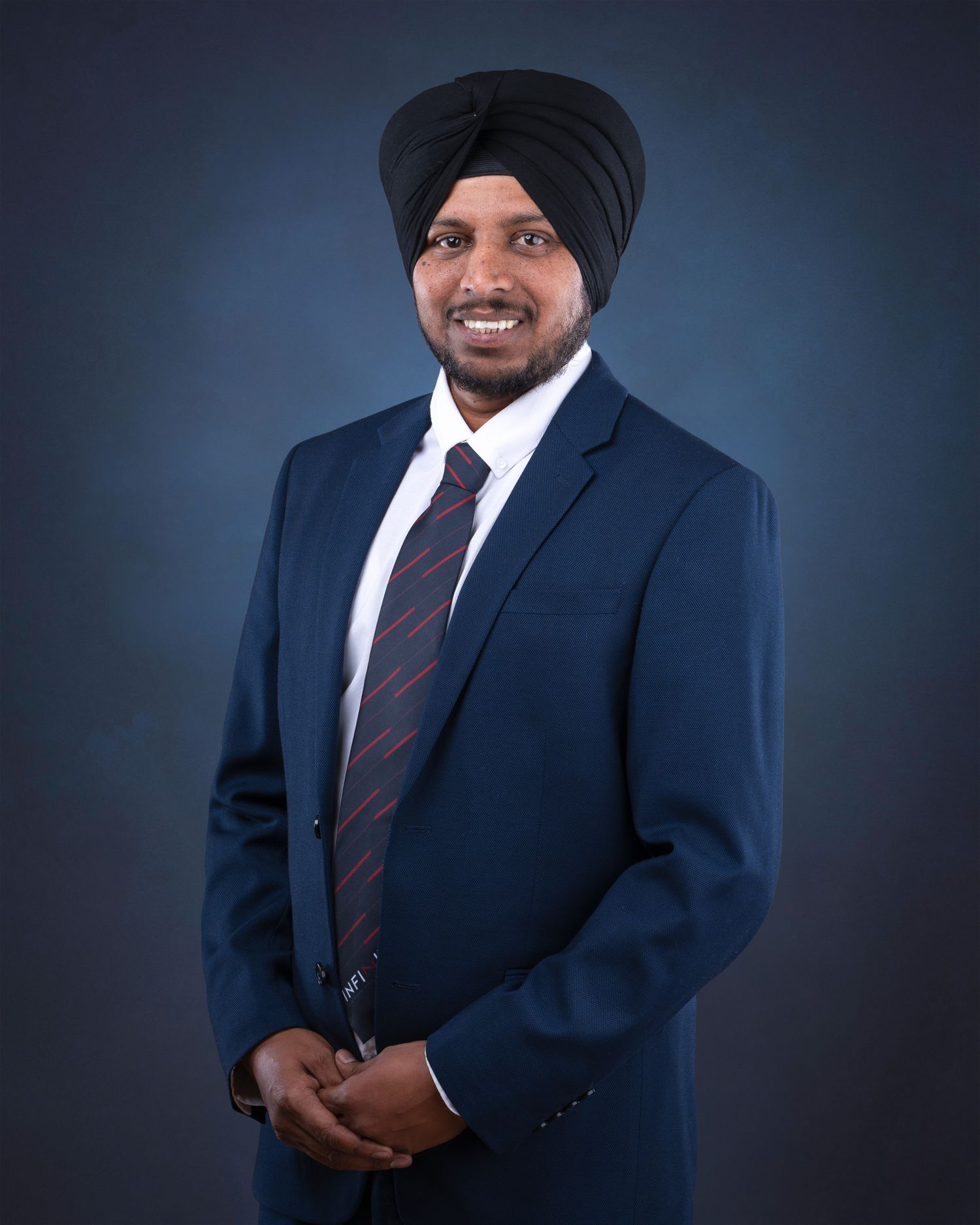 Professional portrait of someone wearing a dark blue suit with a striped tie and black turban against a dark background.