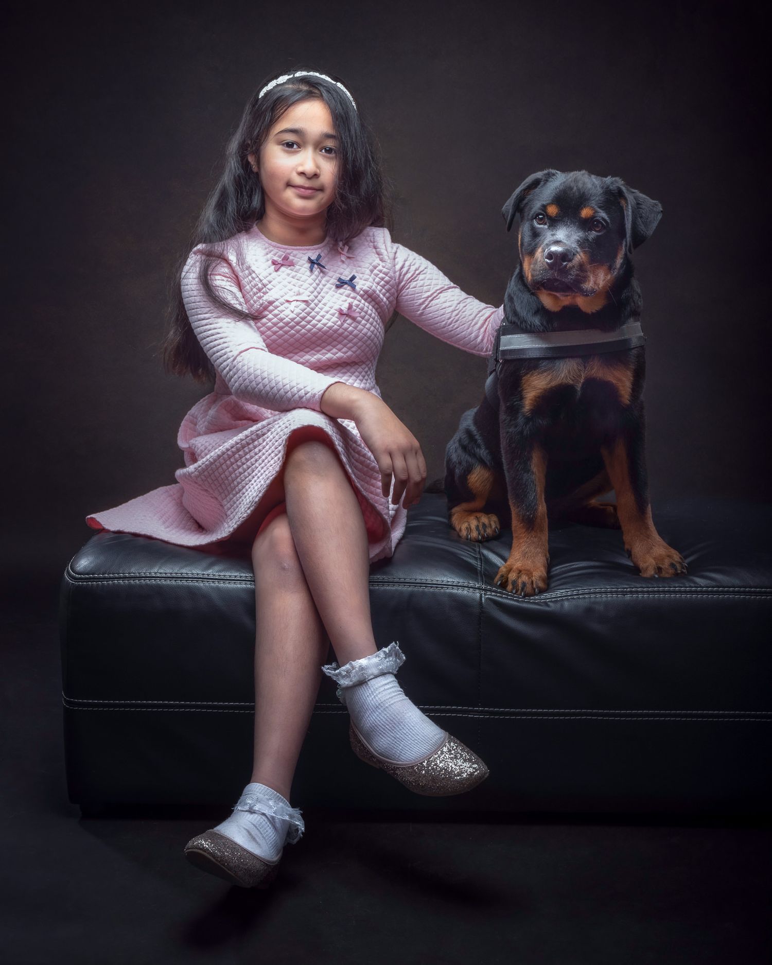 A girl in a pink dress sits on a black leather bench with a Rottweiler dog in a moody studio portrait setting.