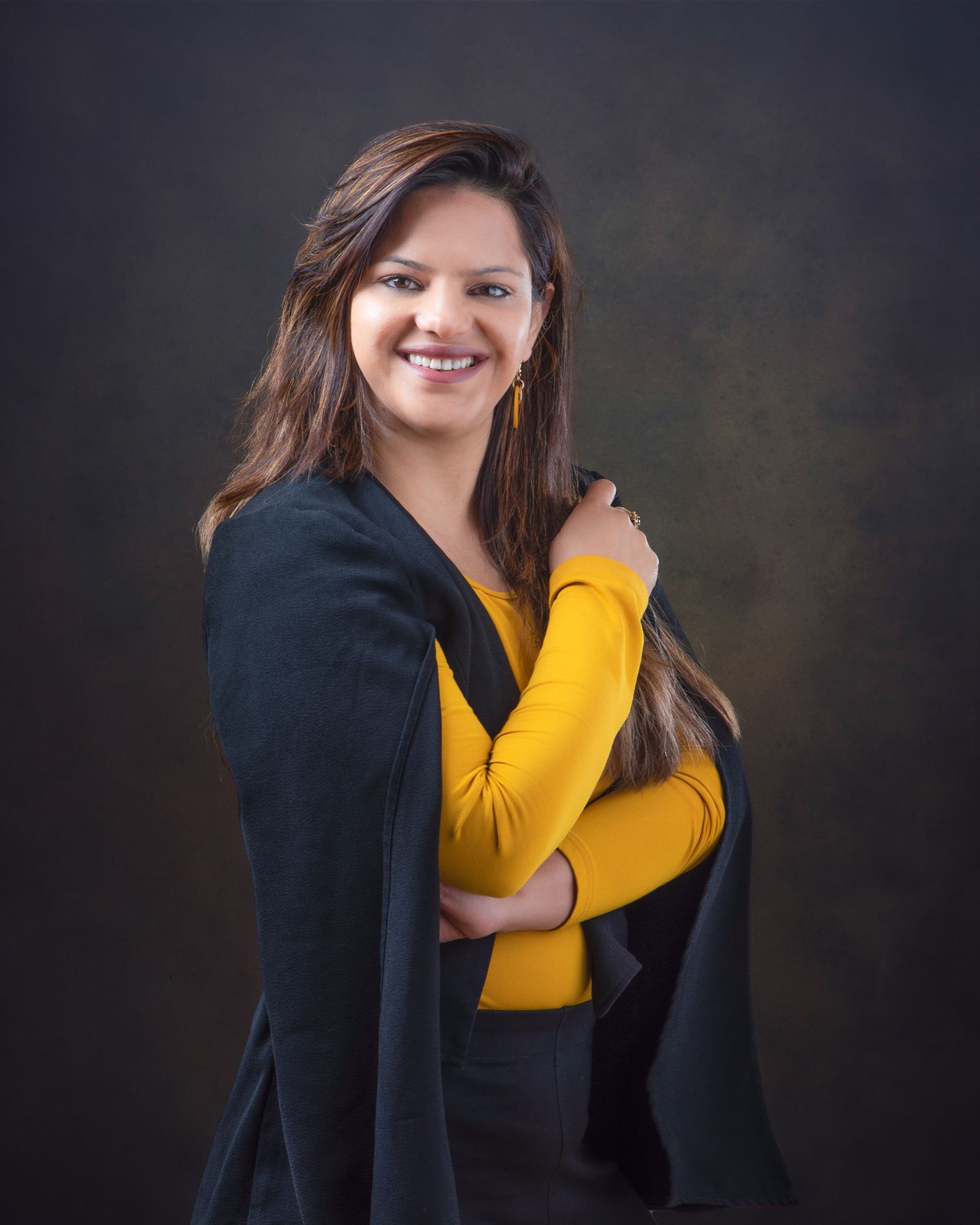 Professional studio portrait against dark background with person wearing yellow top and black blazer posing confidently.