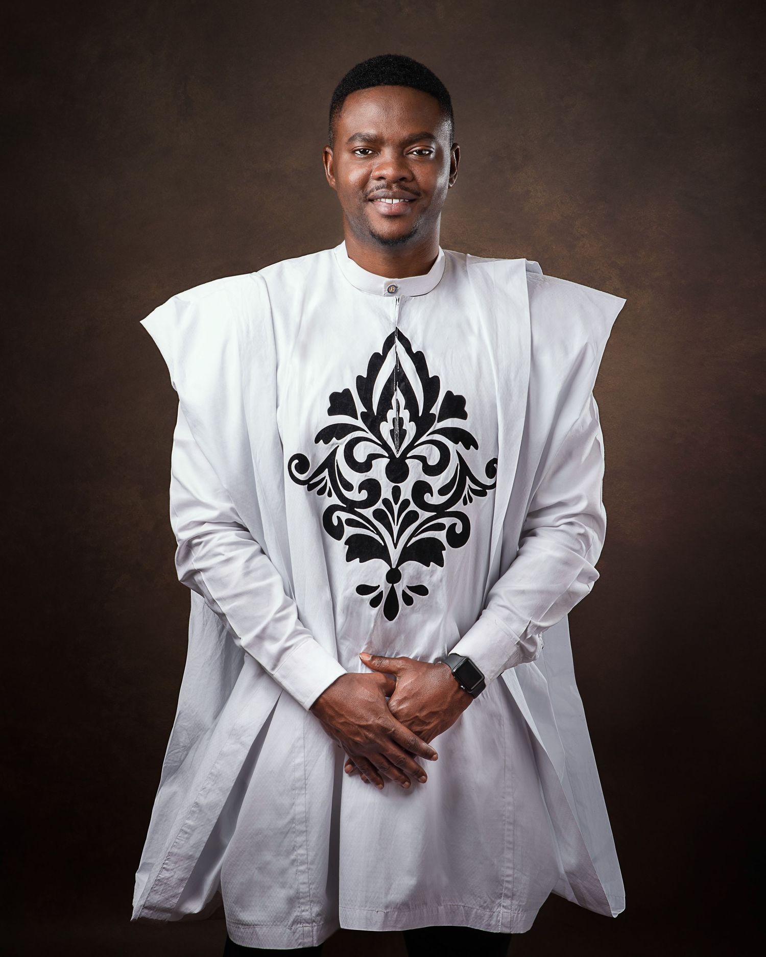Person wearing a white traditional outfit with black damask pattern standing against dark background.