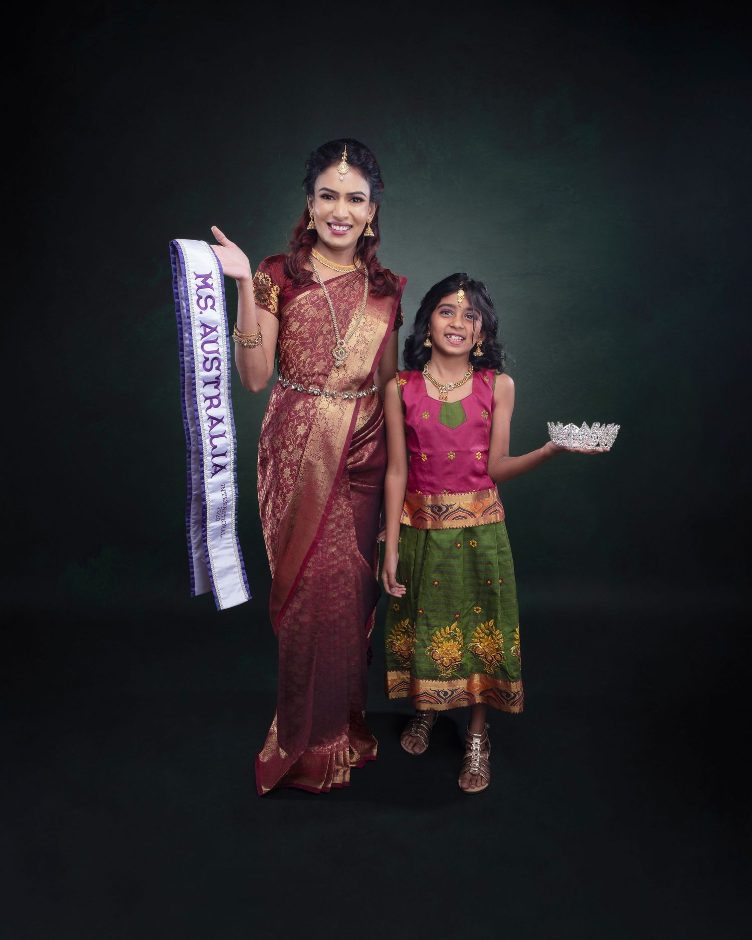 Two people in traditional Indian attire pose against a dark background, one wearing a sari and pageant sash and another in colorful dress.