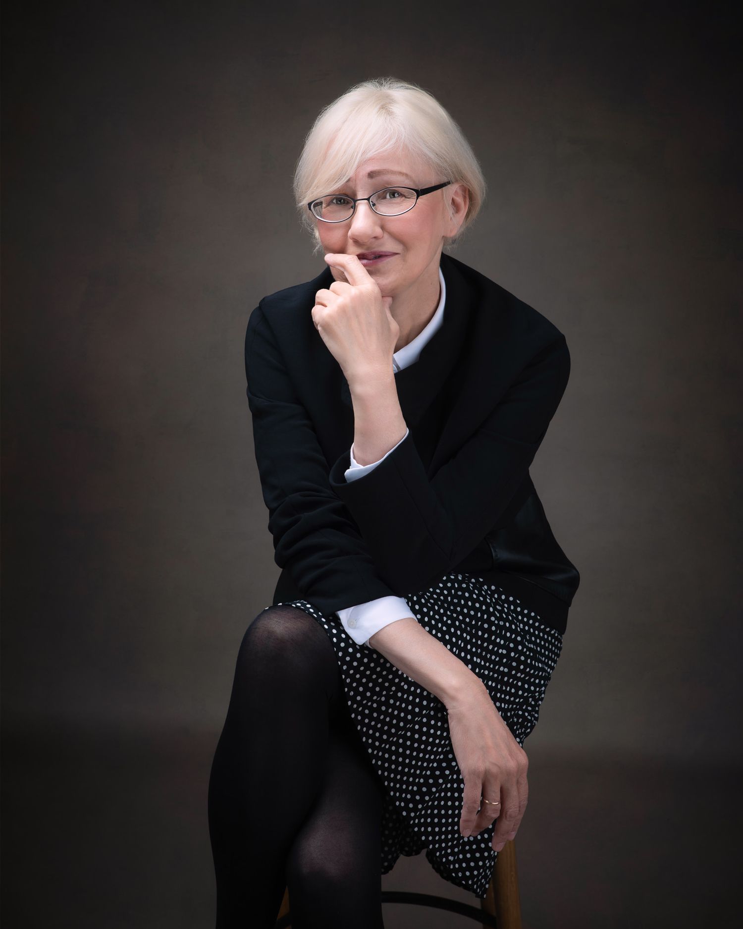 Portrait of someone in a black sweater and polka dot skirt sitting thoughtfully against a dark background.