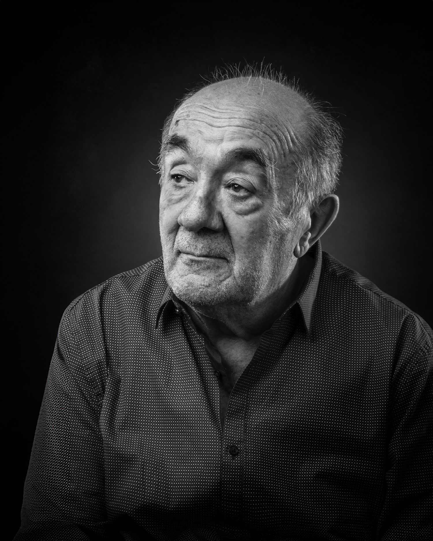 A dramatic black and white portrait photograph of an elderly man in a dark shirt looking contemplative against dark background.