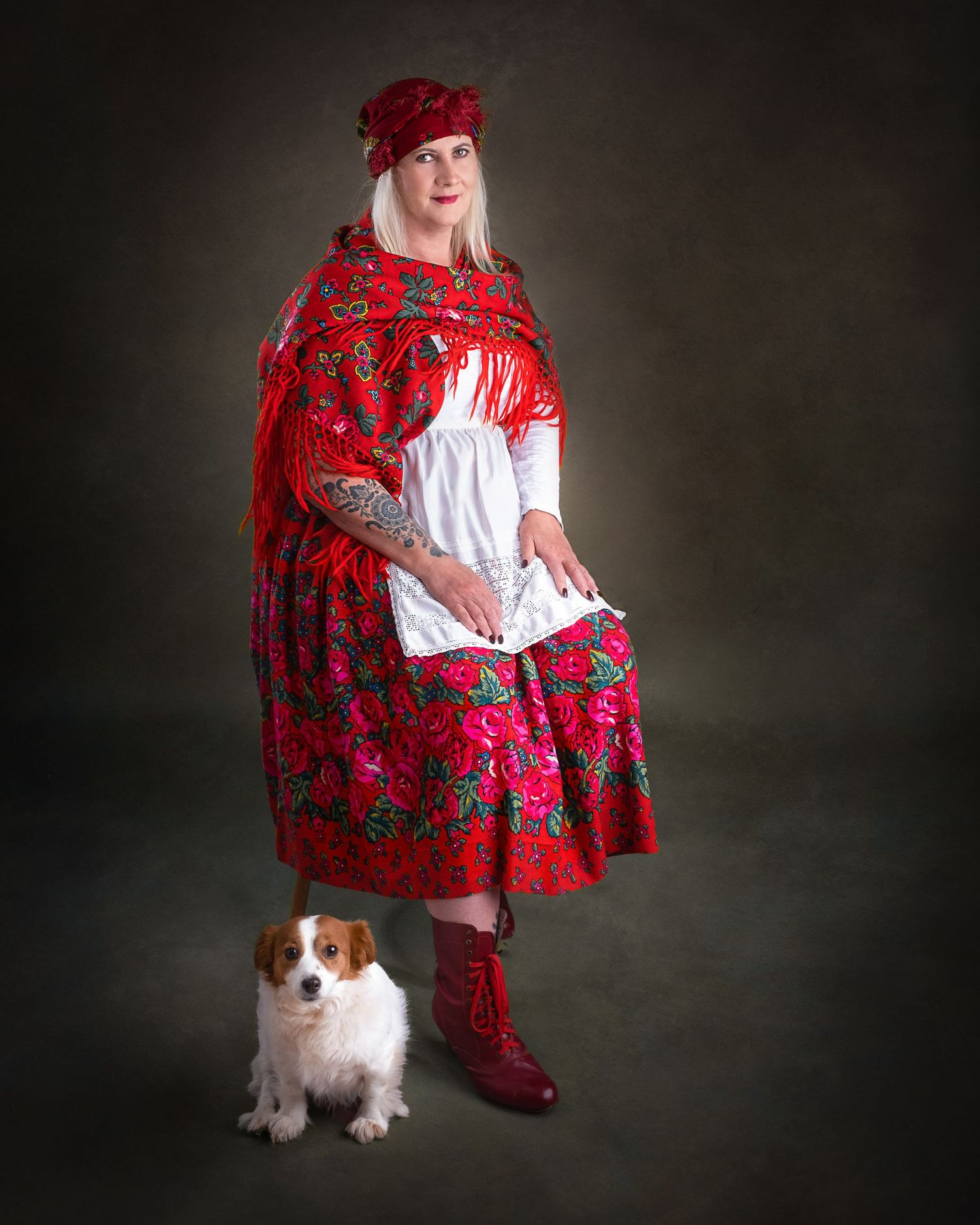 A figure in traditional red folk dress and matching hat stands with a small brown and white dog against a dark backdrop.